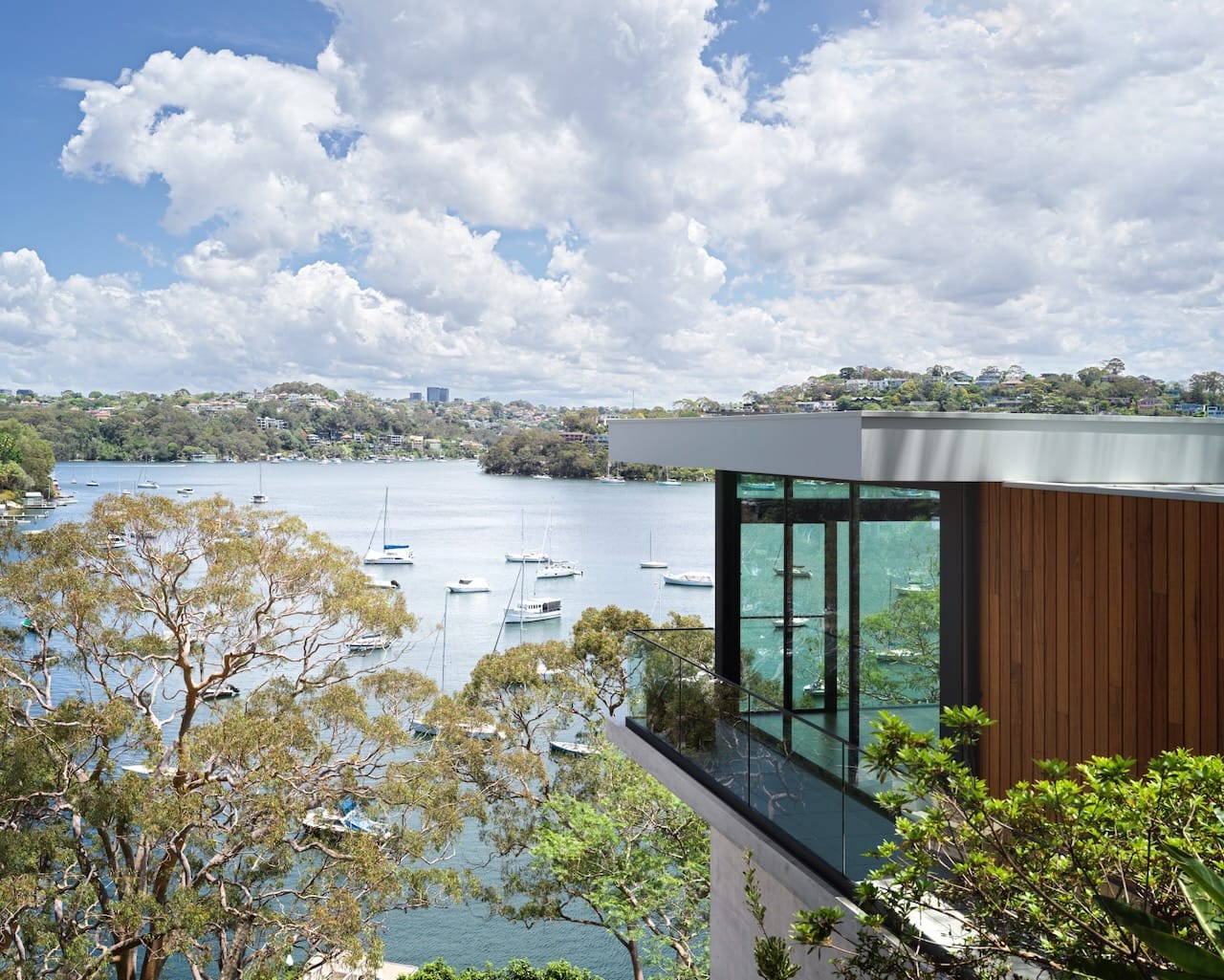 Corbel House by Nick Bell Architects. Photography by Justin Alexander. Panoramic views over harbour and hilly bushland, with timber and glass double story home in right bottom corner. 