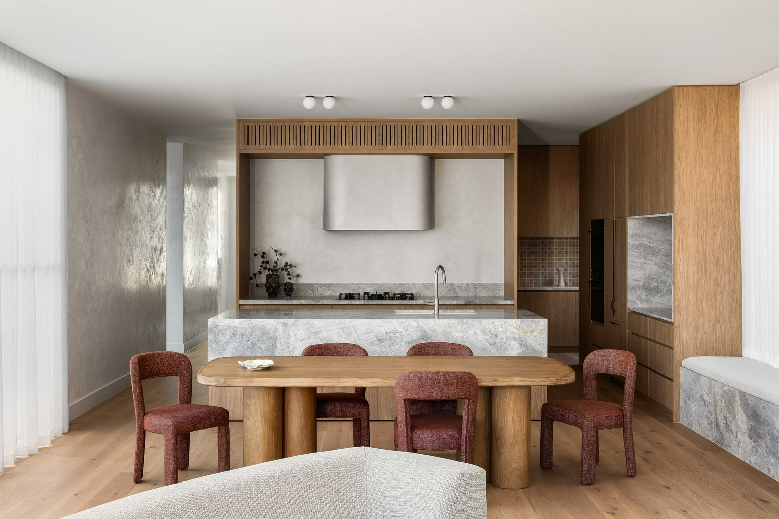 King House by Taouk Architects. Photography by Emily Bartlett. Open plan kitchen and dining area with grey marbled island, timber cabinetry, flooring and dining table with terracotta boucle chairs. 