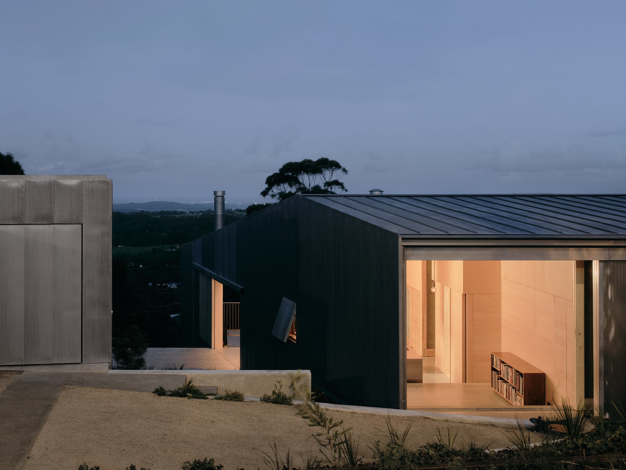 Mapleton House by Atelier Chen Hung. Photography by David Chatfield. Large opening in facade of home, showing simple interior with a bookshelf and warm lighting. 
