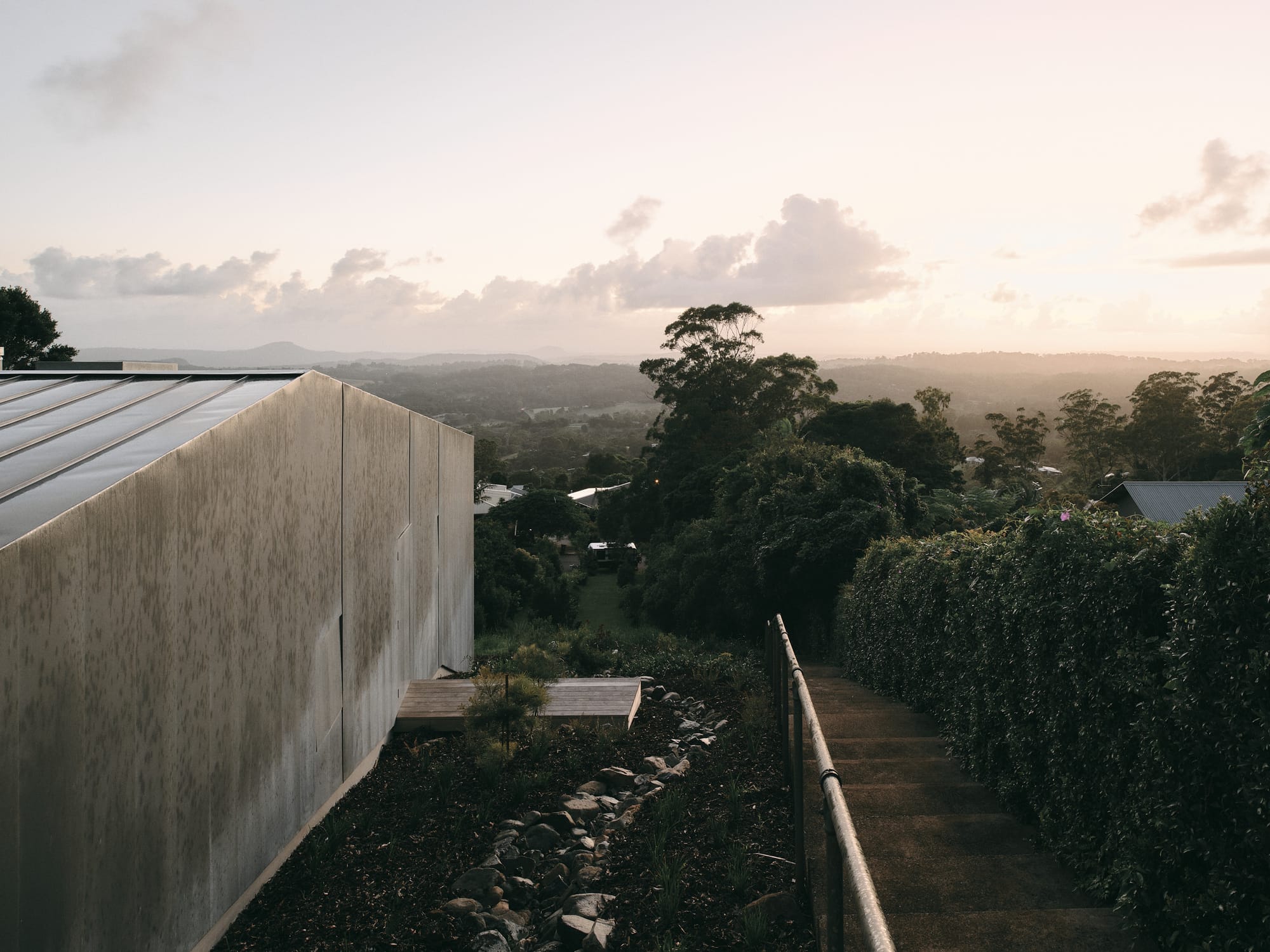 Mapleton House by Atelier Chen Hung. Photography by David Chatfield. Rolling landscape with hills, dense foliage along exterior staircase and a zinc-clad structure to the left. 