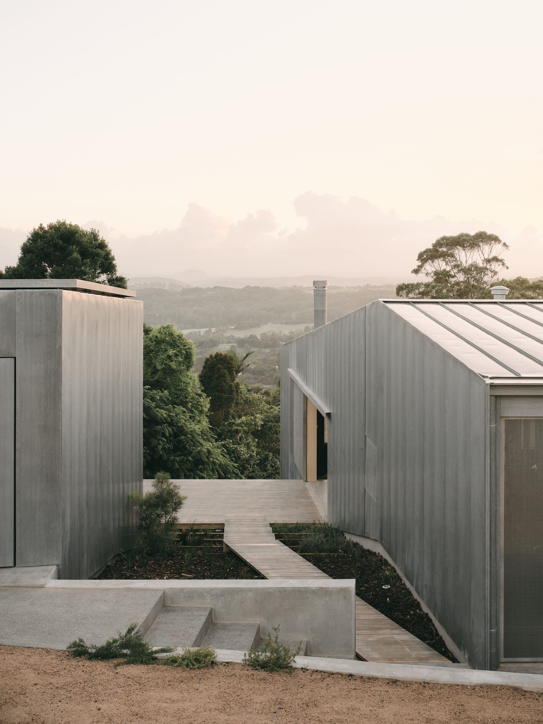 Mapleton House by Atelier Chen Hung. Photography by David Chatfield. Views over valley, with zinc-clad home, deck and concrete steps in foreground. 