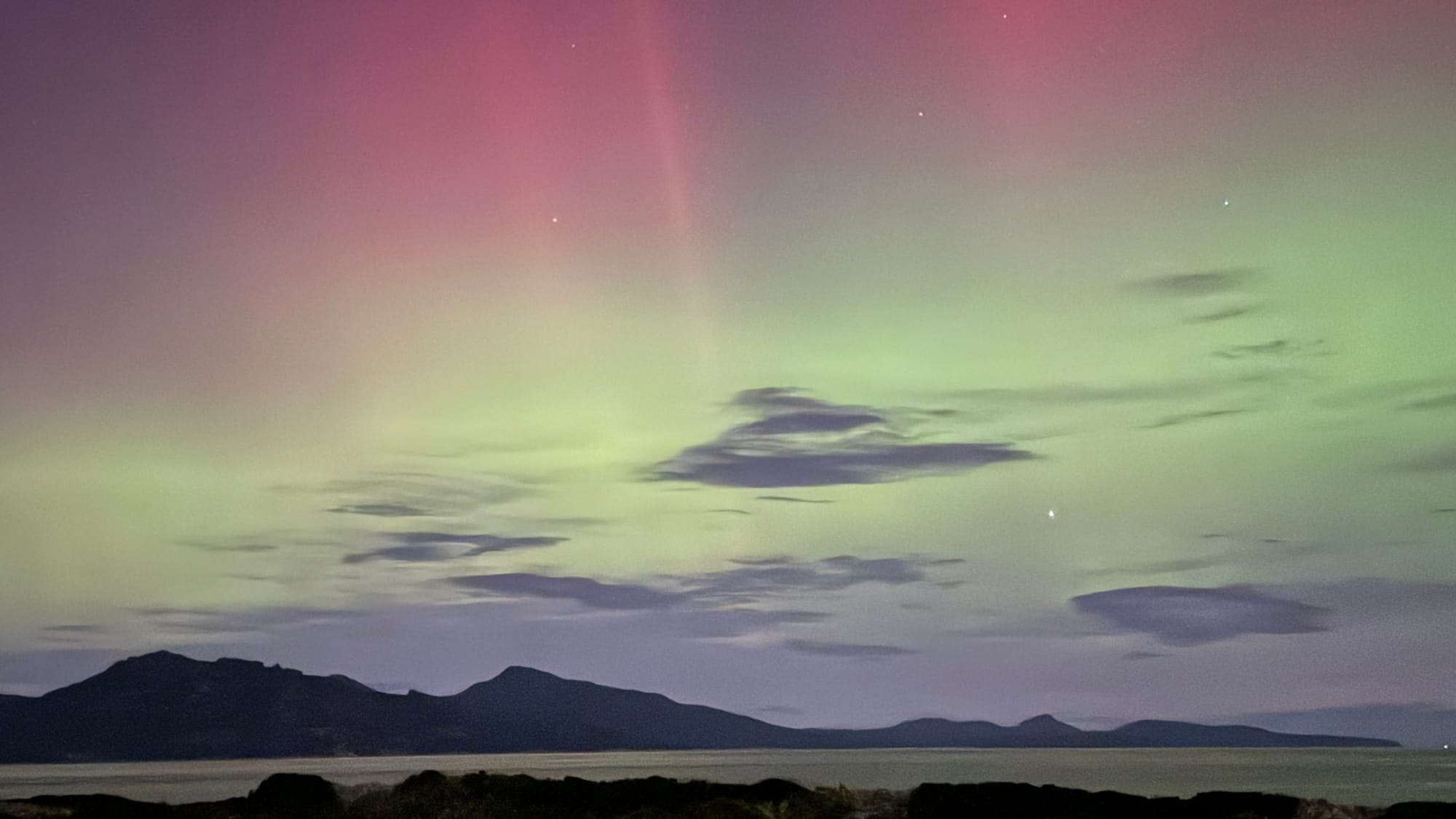 Aurora Australia at Studio Tasmania. Photography by Studio Tasmania. Aurora with pink and teal tones over ocean and hilly landscape in background. 