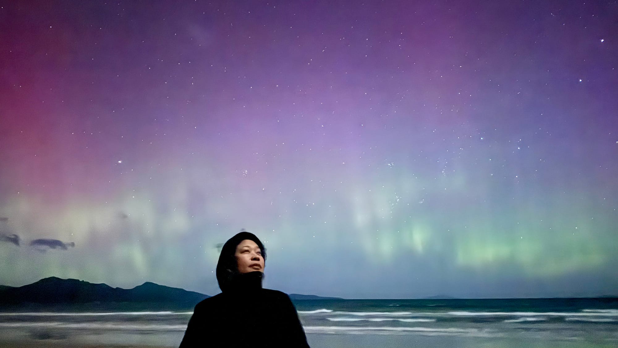 Aurora Australia at Studio Tasmania. Photography by Studio Tasmania. Aurora in pink, purple and teal hues behind woman in black windbreaker. 