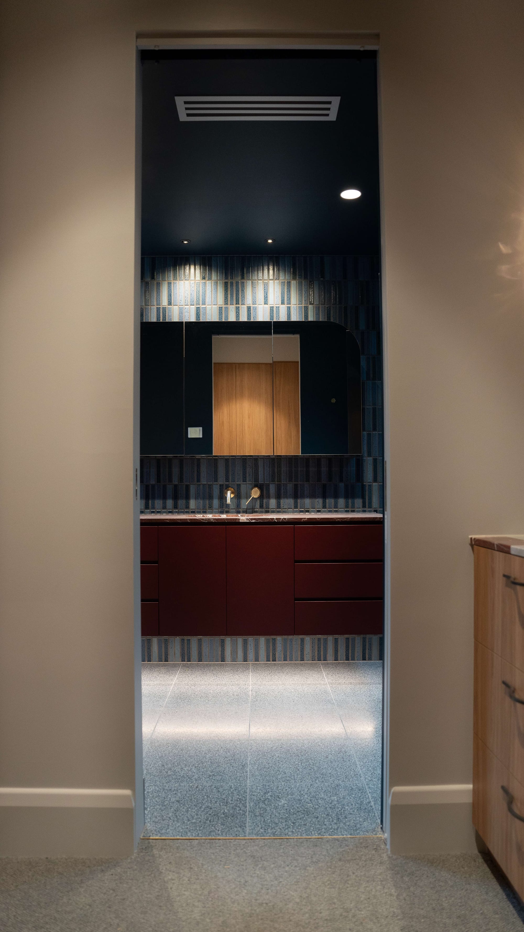 Murrumbeena by PREM.BUILT. Image copyright of Prem.Built. Doorway framing bathroom with blue floor tiles, blue mosaic wall tiles, burgundy cabinetry and mirrored storage. 