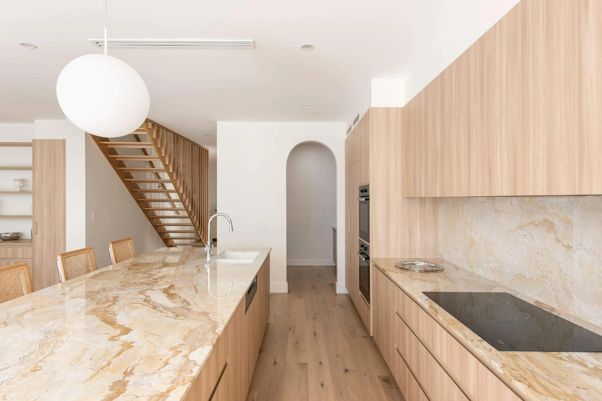 MCCA House by RADS. Kitchen with arched entry, beige marble countertops, timber cabinetry and flooring.