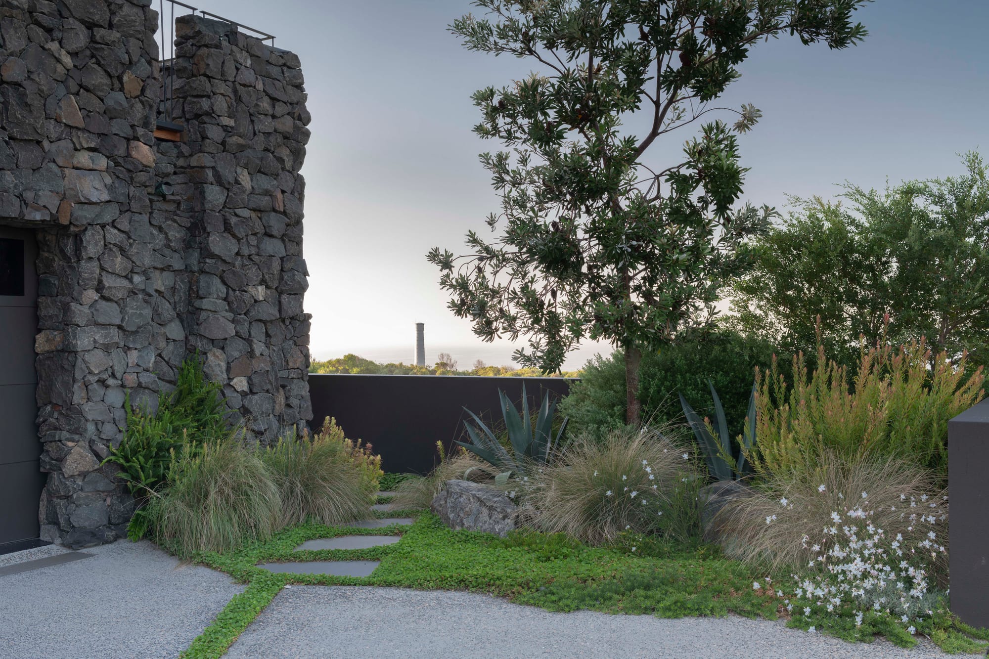 Stone House by Secret Gardens.Entry path edged with bluestone and coastal planting leading toward a stone wall.