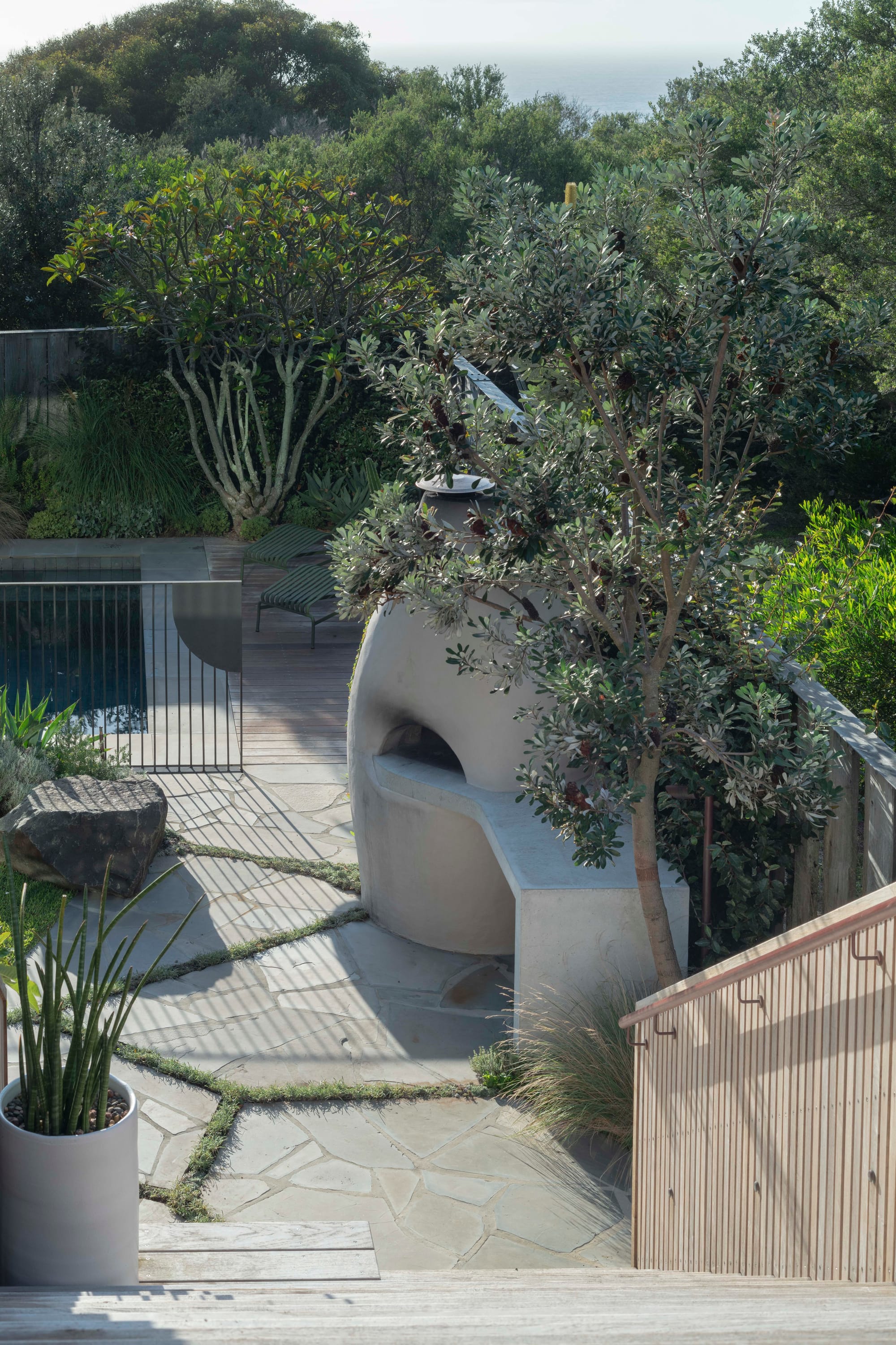 Stone House by Secret Gardens. Curved outdoor pizza oven set within a bluestone terrace surrounded by trees and layered coastal planting.