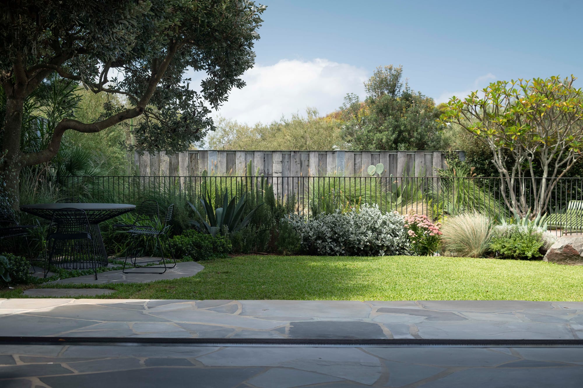 Stone House by Secret Gardens. Garden view across bluestone paving toward the pool, framed by native shrubs and flowering plants.