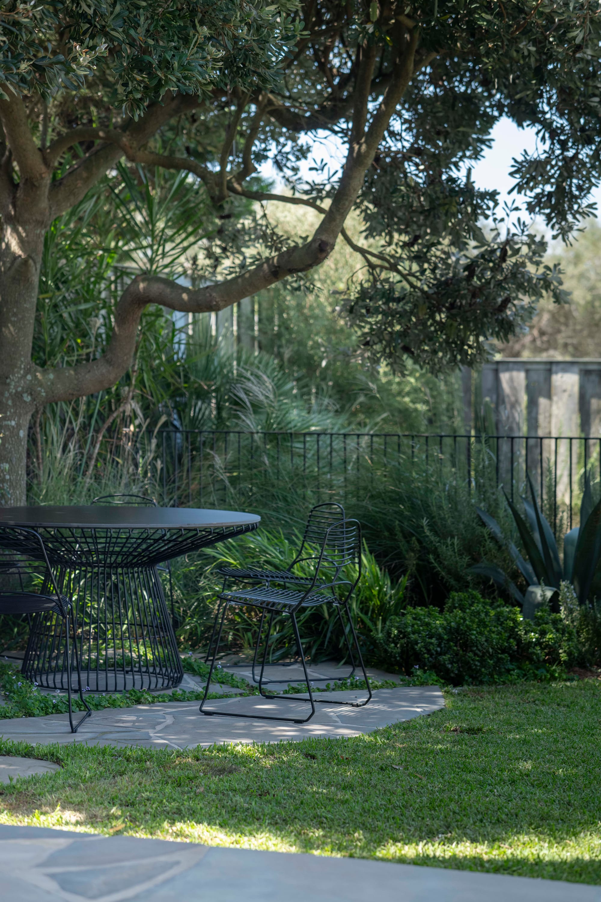 Stone House by Secret Gardens. Shaded outdoor dining area beneath a mature tree with black wire furniture and stone paving.