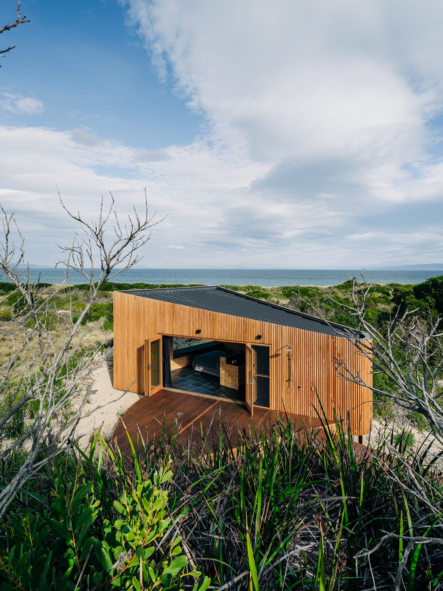 Studio Tasmania. Photography by Adam Gibson. Timbe clad beach hut with timber deck, surrounded by beach foliage. 