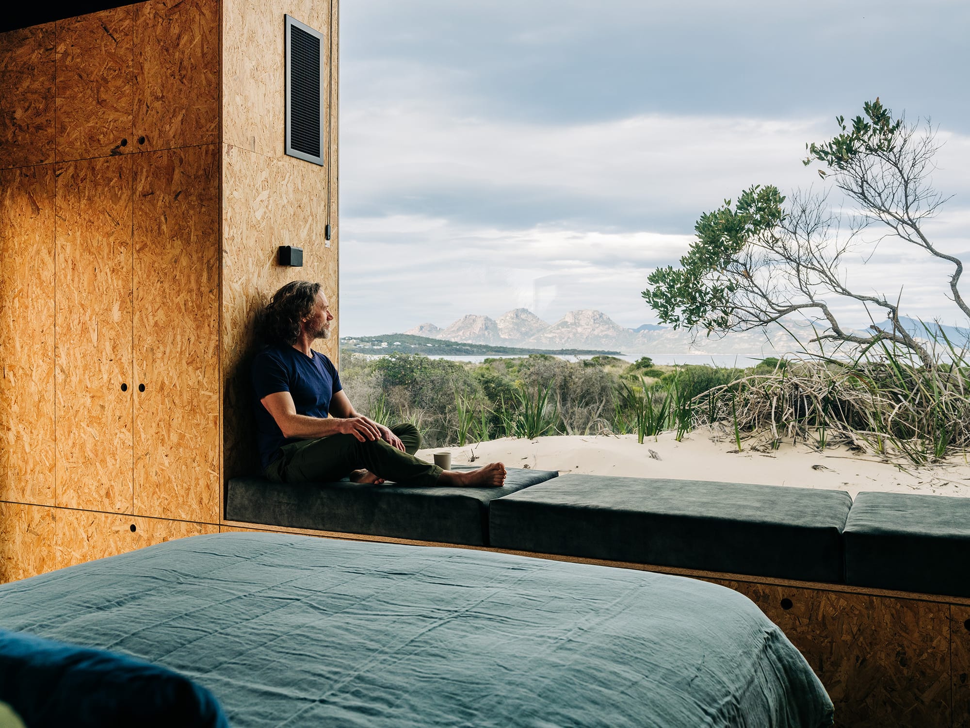 Studio Tasmania. Photography by Adam Gibson. Man sitting on window seat overlooking sand dunes, plants and hills in distance. 
