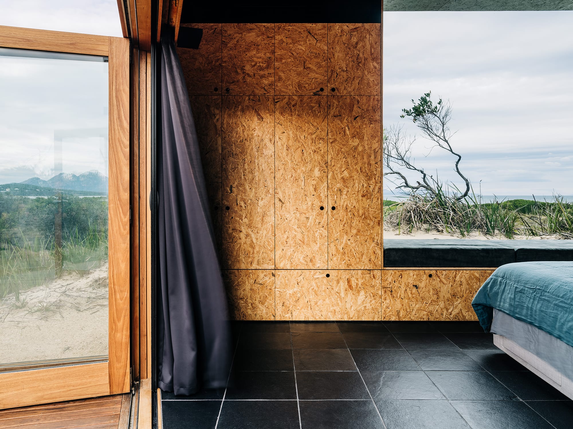 Studio Tasmania. Photography by Adam Gibson. Bedroom space with black tiled floors, timber walls and cabinetry, blue bedding and large glass windows and doors with views to beach. 
