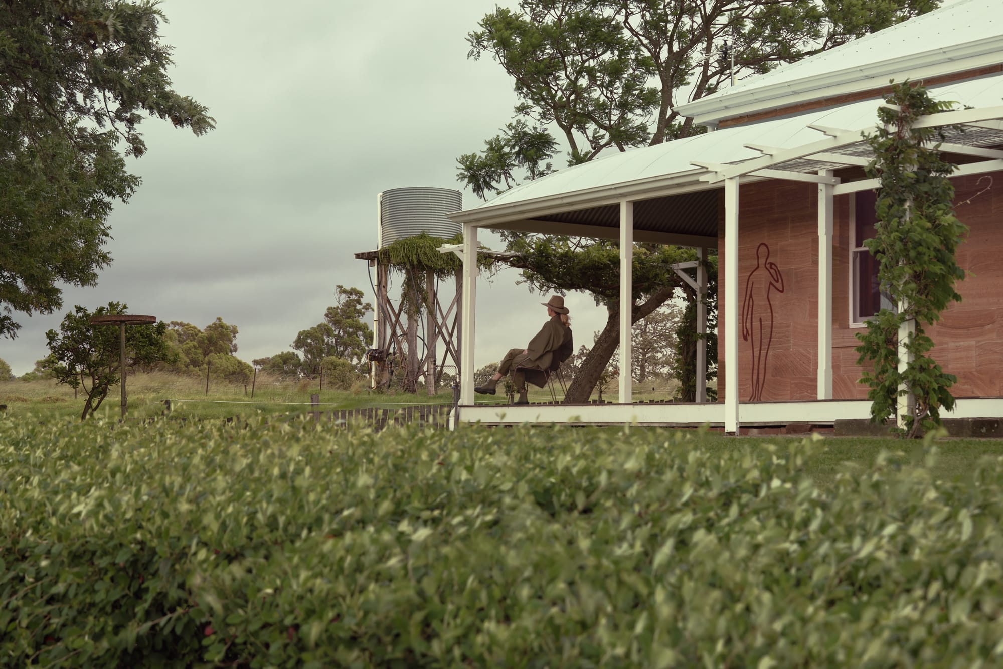 Binnum Homestead by Alexandra Buchanan Architecture. Photography by Cieran Murphy. Woman sitting on deck wrapping around traditional sandstone homestead, with water tower in background. 