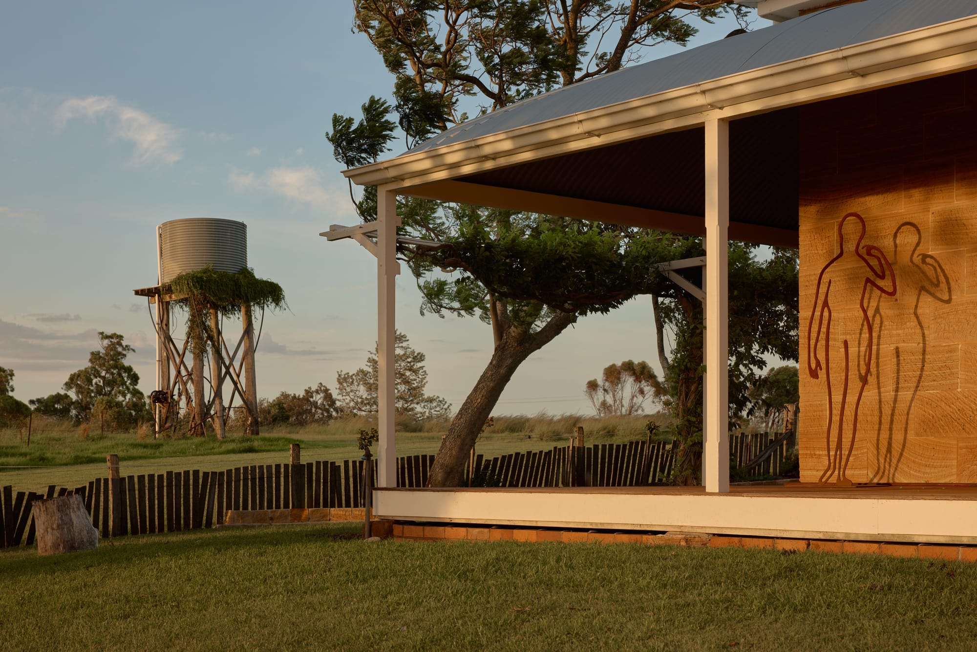 Binnum Homestead by Alexandra Buchanan Architecture. Photography by Cieran Murphy. Sadnstone homestead structure with wraparound timber deck, grassed yard and watertower in background. 
