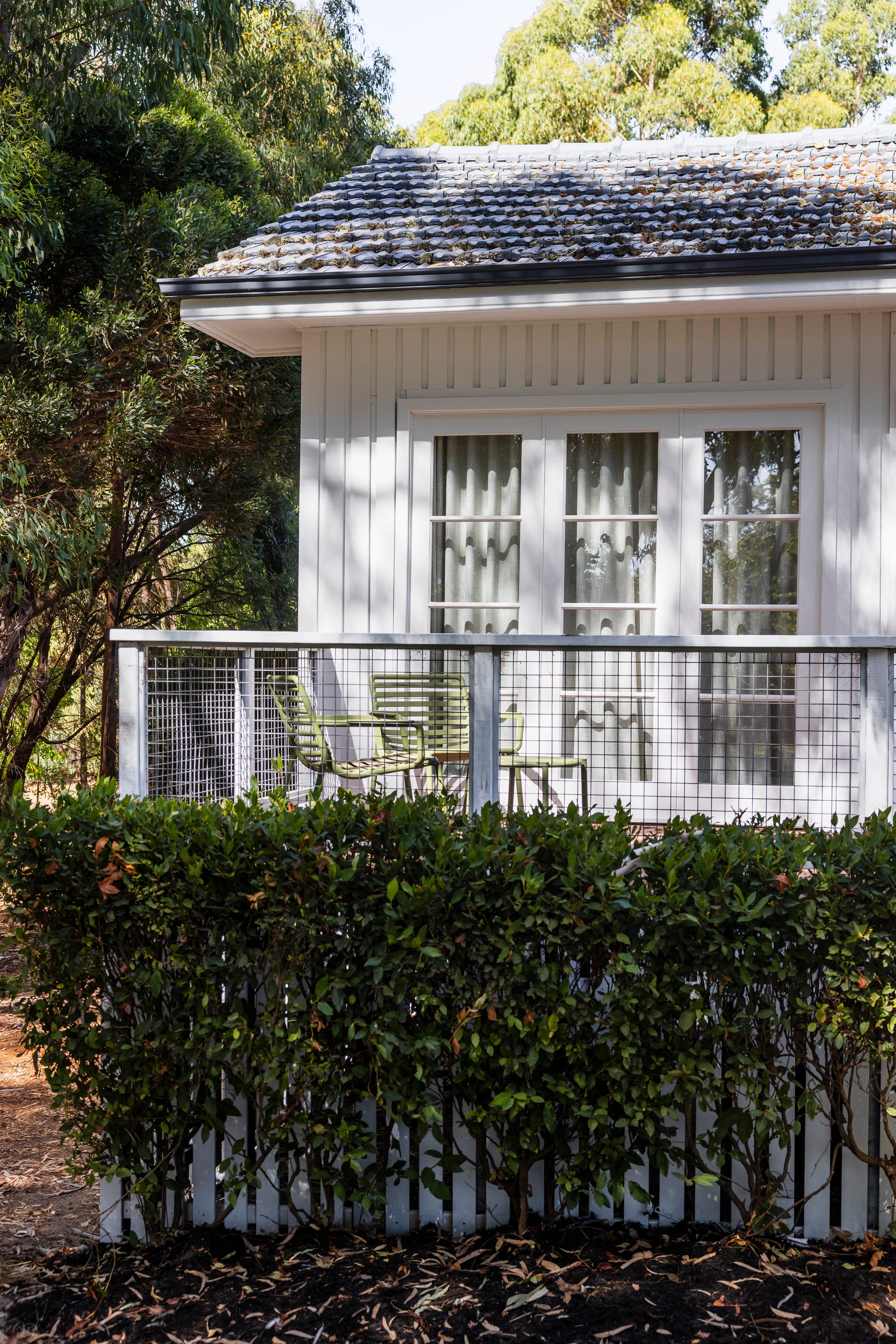 The Cottage by Bel.Bon Design. Photography by Sana Brotherson. Rear facade of white timber clad cottage with green dining seating on verandah, and picket fence with low hedge. 