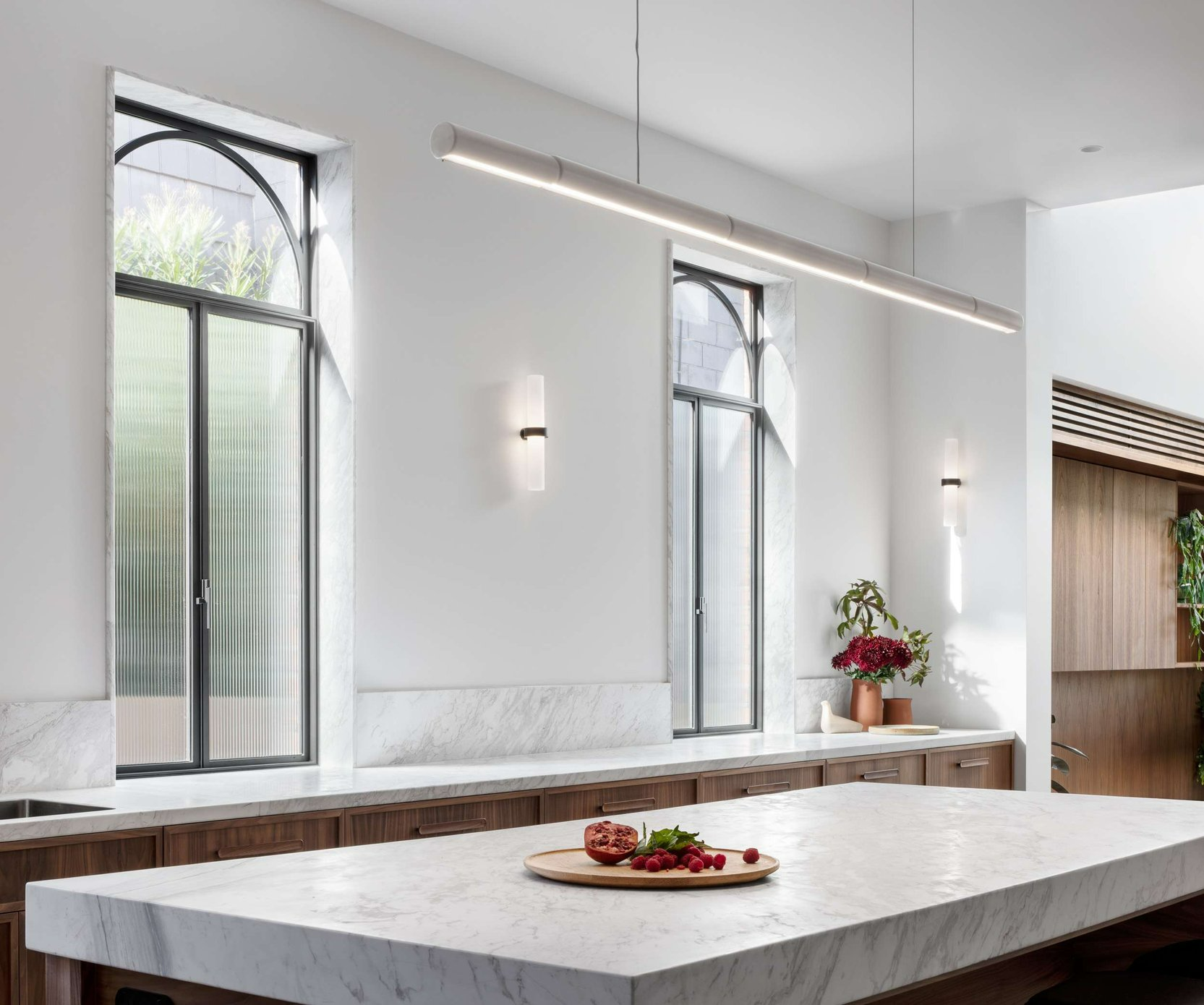 South Terrace Renovation by Chan Architecture. Photography by Tatjana Plitt. Kitchen counter with white stone countertop and half-height splashback, timber cabinets, fluted glass, black framed arched windows and white walls.
