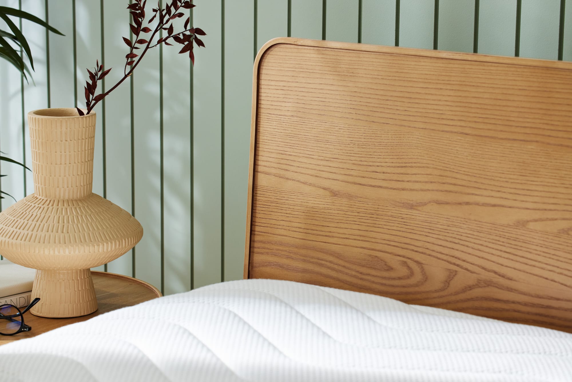 Kirribilli Bed Frame by Koala. Close up of timber headboard in front of sage wood paneled walls and beige abstract vase on side table.