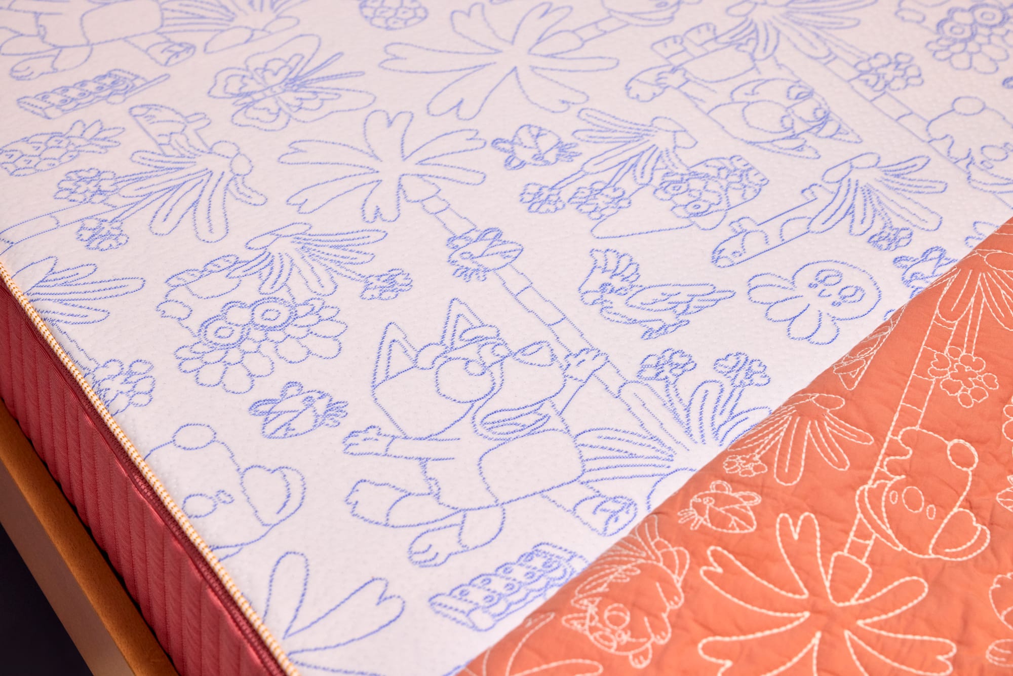 Koala x Bluey Snoozytime Mattress by Koala. Close-up of white and orange mattress with white and blue stitched characters and plants. 