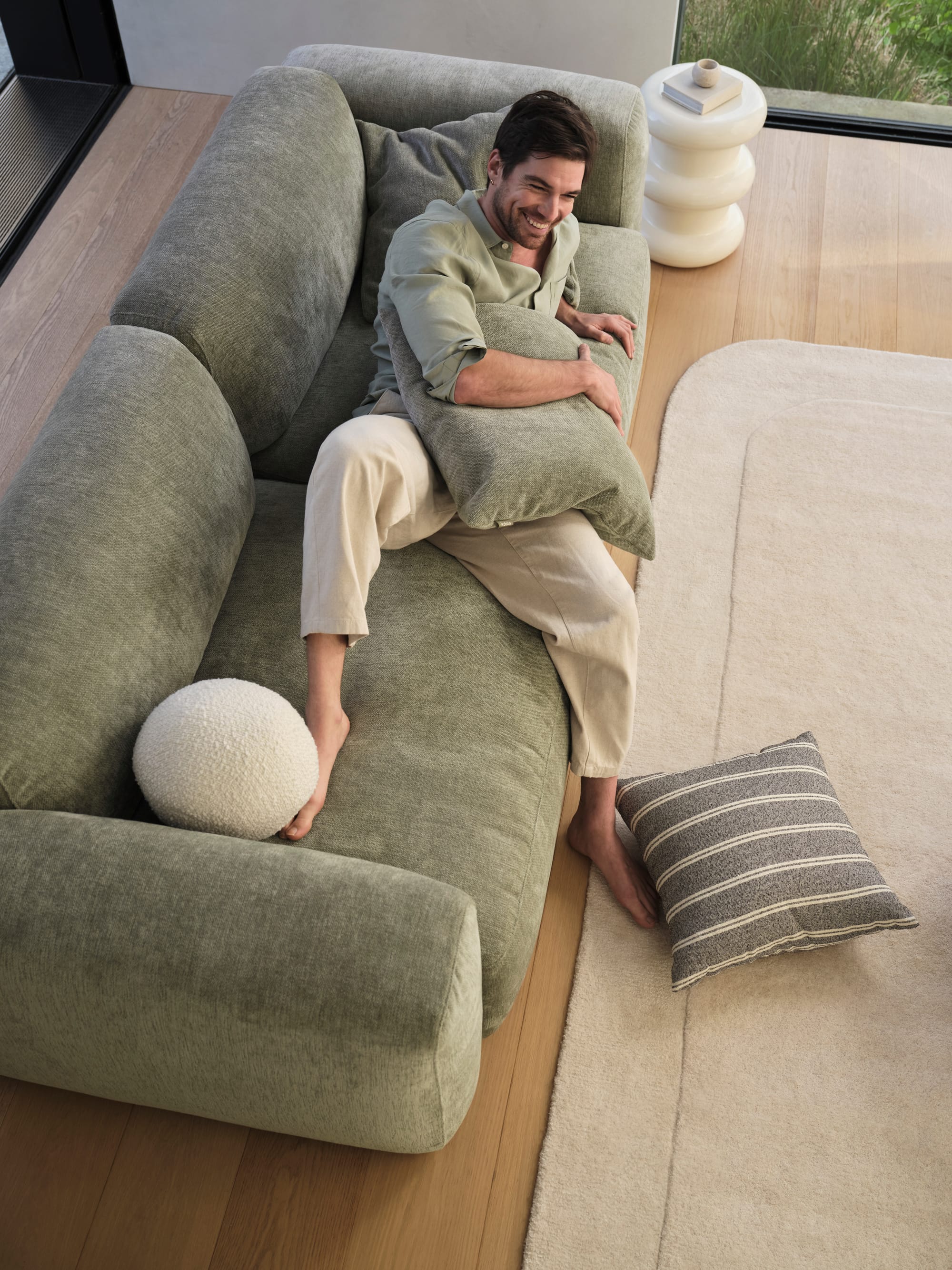 Torquay Sofa in Forest Dawn by Koala. Birds-eye-view of man lounging on green modular lounge, wearing green shirt in room with timber floors and white rug. 