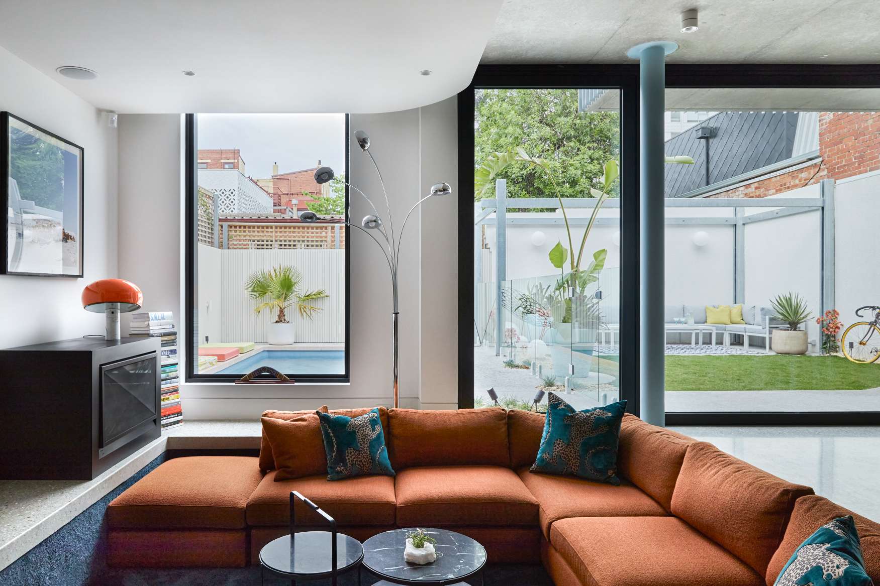 Park St by SGKS ARCH. Photography by Tania Lee. Orange sunken lounge with teal throw cushions, in living space with white tiles and black windows overlooking backyard. 