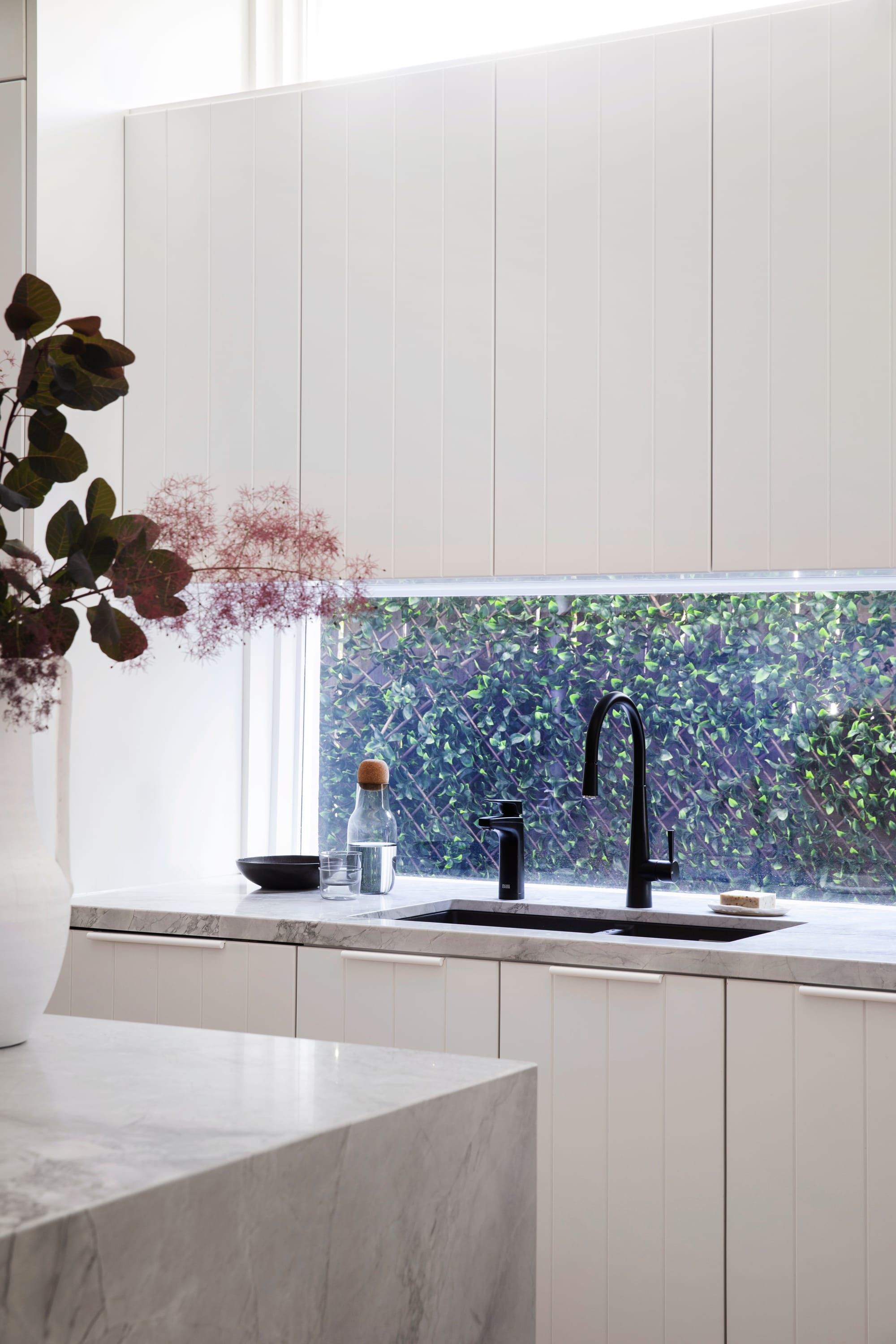 Bondi Beach House by Baikie Corr Architecture. Photography by Chris Warnes. Kitchen with white cabinets, stone countertops, glass window splashback looking over garden, and black fixtures. 
