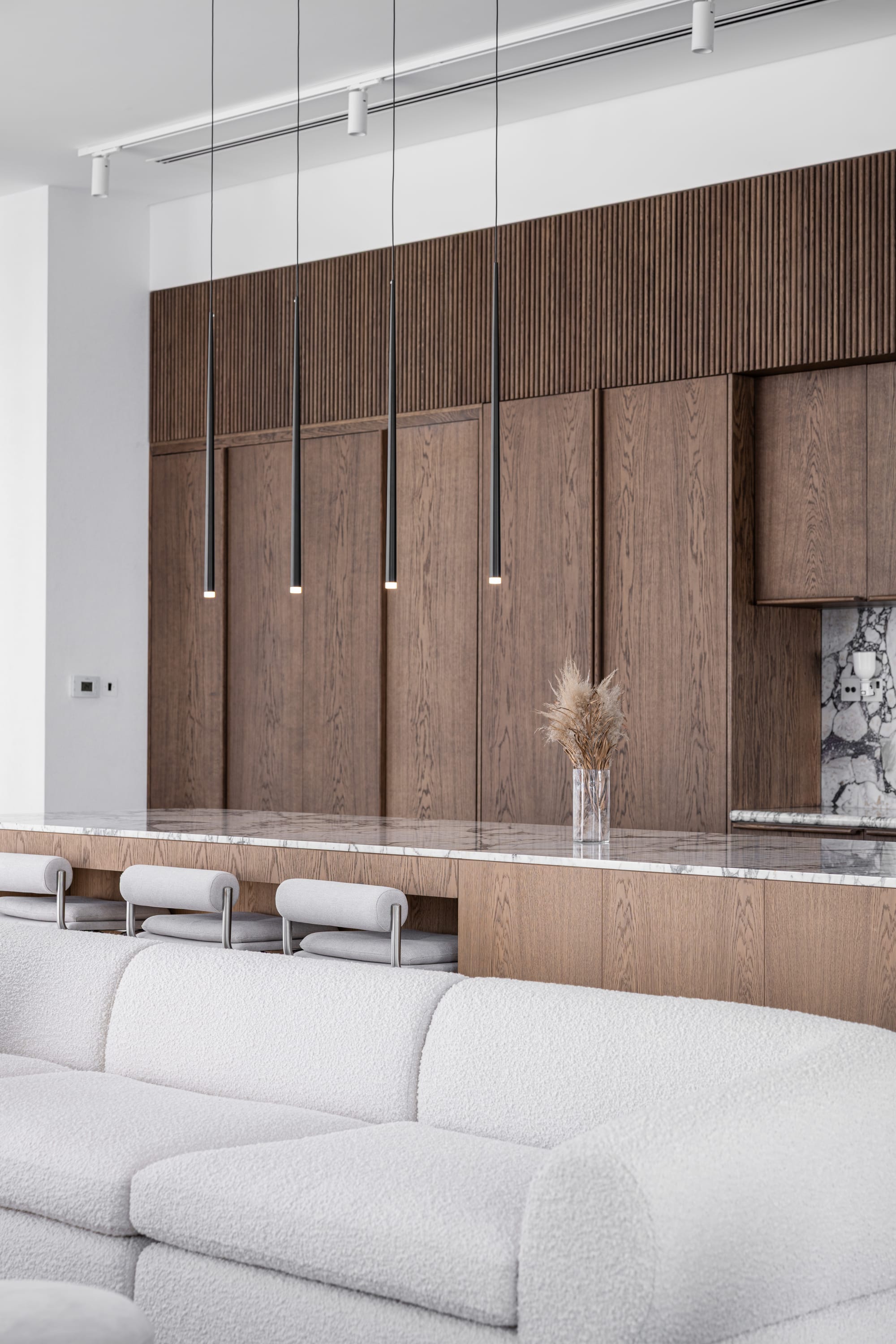 Veil House by ENCLAVE Architects. Photography by Hassan Arante & Gener Rayes. Kitchen with timber cabinetry, stone splashback and countertops, behind a white boucle sofa. 