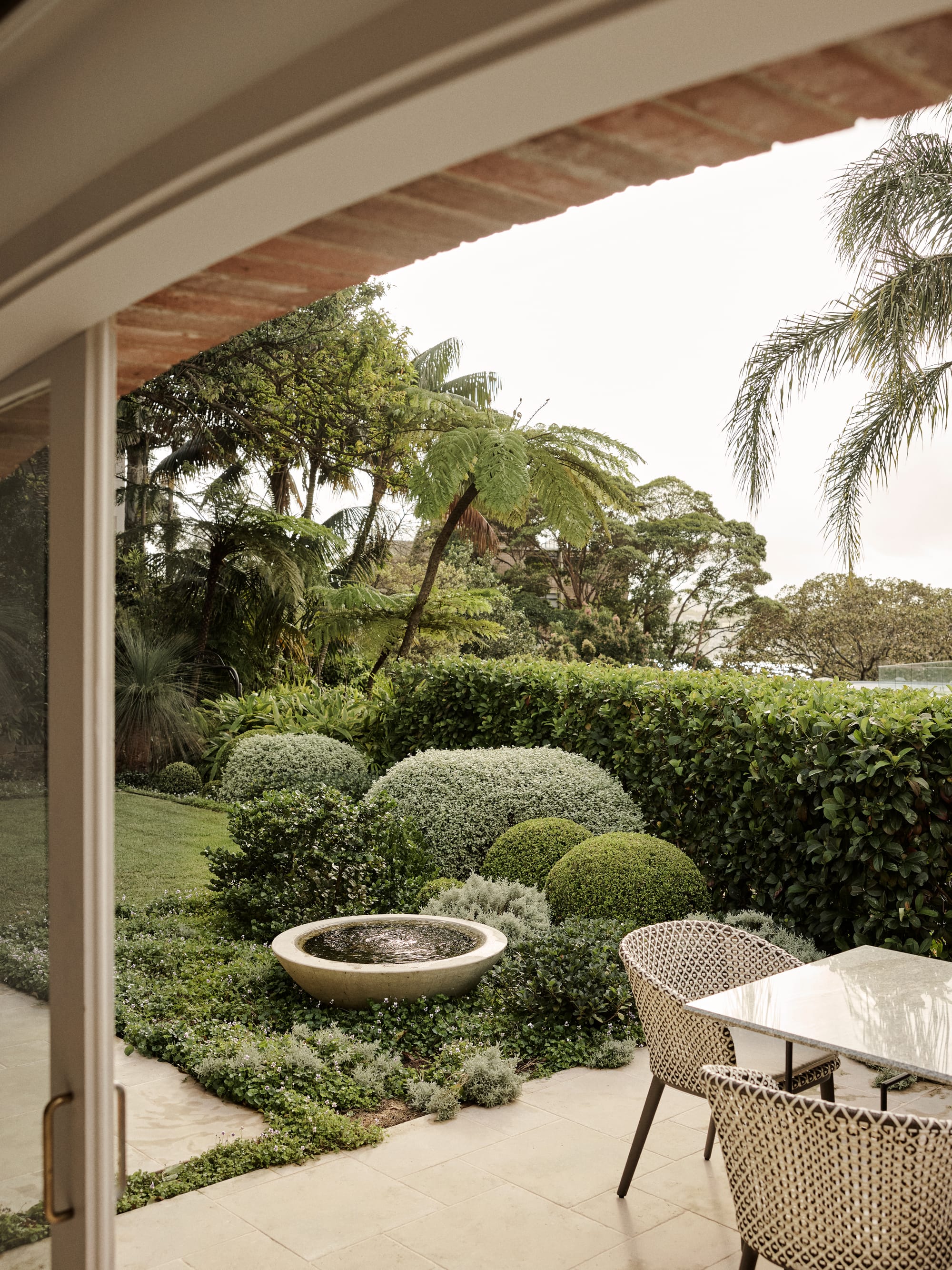 The Darling Edit by Marylou Sobel Interior Design. Photography by Anson Smart. Garden with paved area with outdoor dining setting, a round water fountain and lush gardens. 