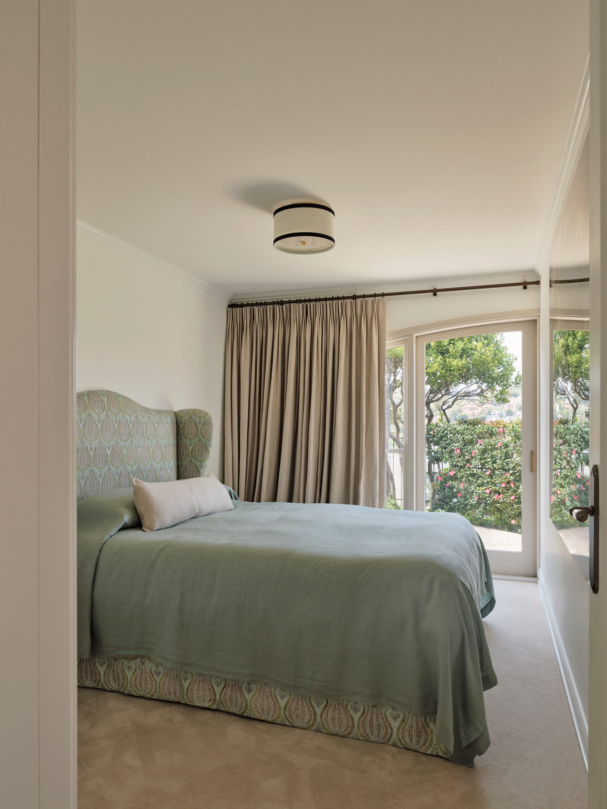 The Darling Edit by Marylou Sobel Interior Design. Photography by Anson Smart. Bedroom with sage patterned fabric headboard, sage linen bedding, beige curtains and glass doors opening onto garden. 