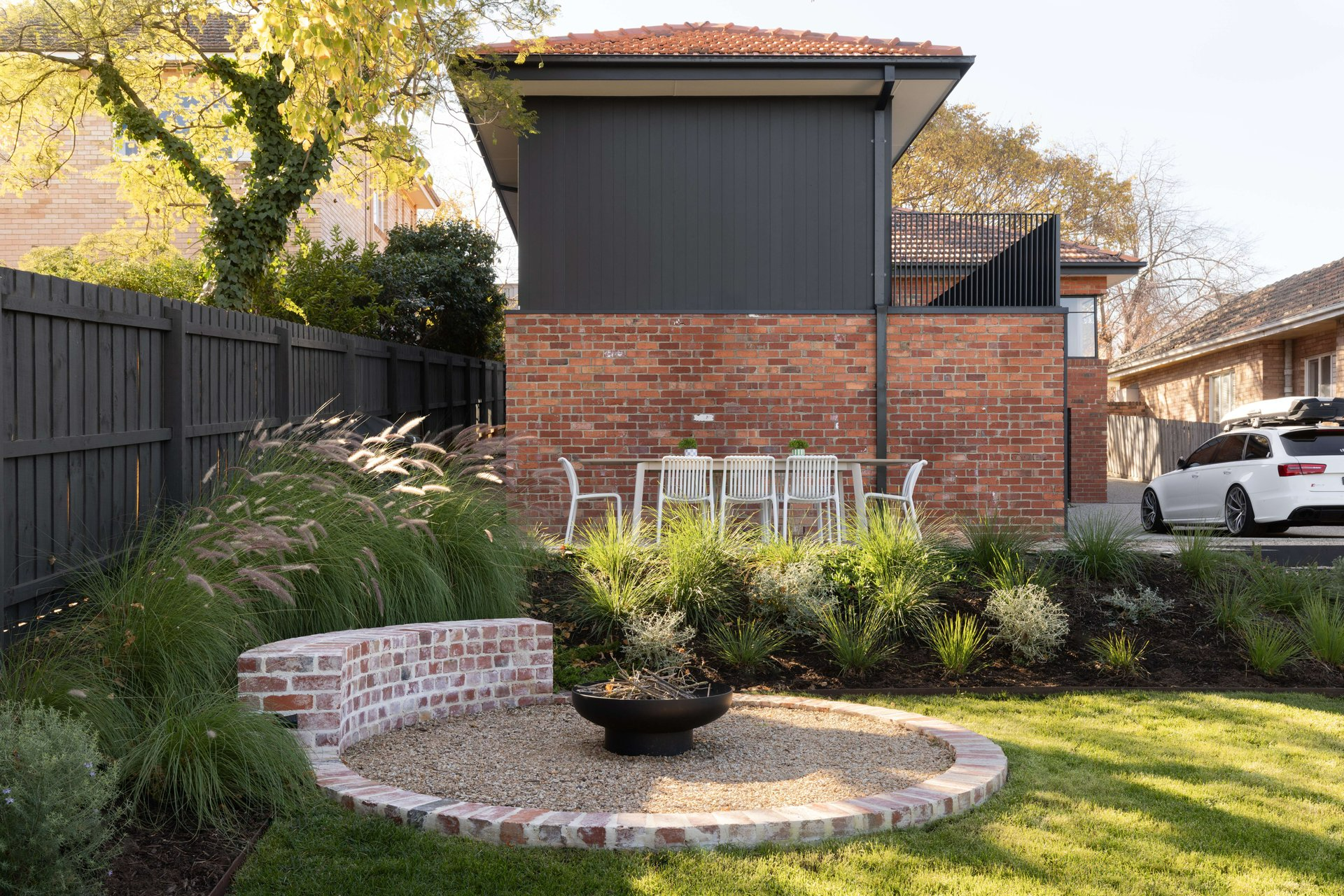 MAUD by Mancini Made. Photography by Elise Scott. Backyad of couble storey red brick and black timber clad home, with recycled brick firepit, low bushes and grass. 