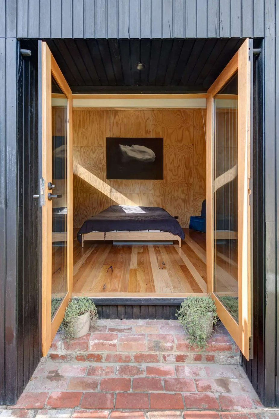 237 York Street, South Melbourne by Plico Design Studio. Photography by Jamie Diaz Berrio. Timber doors opening inwards from paved exterior to interior bedroom with timber flooring, plywood walls and a navy bed. 