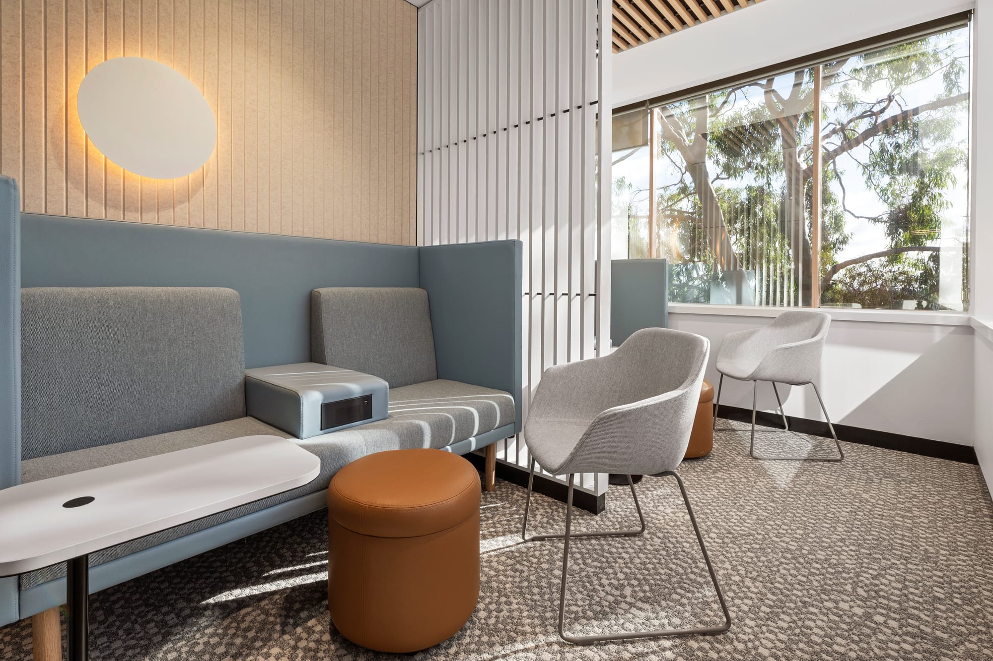 MDA National by Saru Design. Photography by Pete Battye. Integrated bench seating in office space with slate grey cushions and chairs, orange leather stools, white timber screen dividers and patterned grey carpet. 