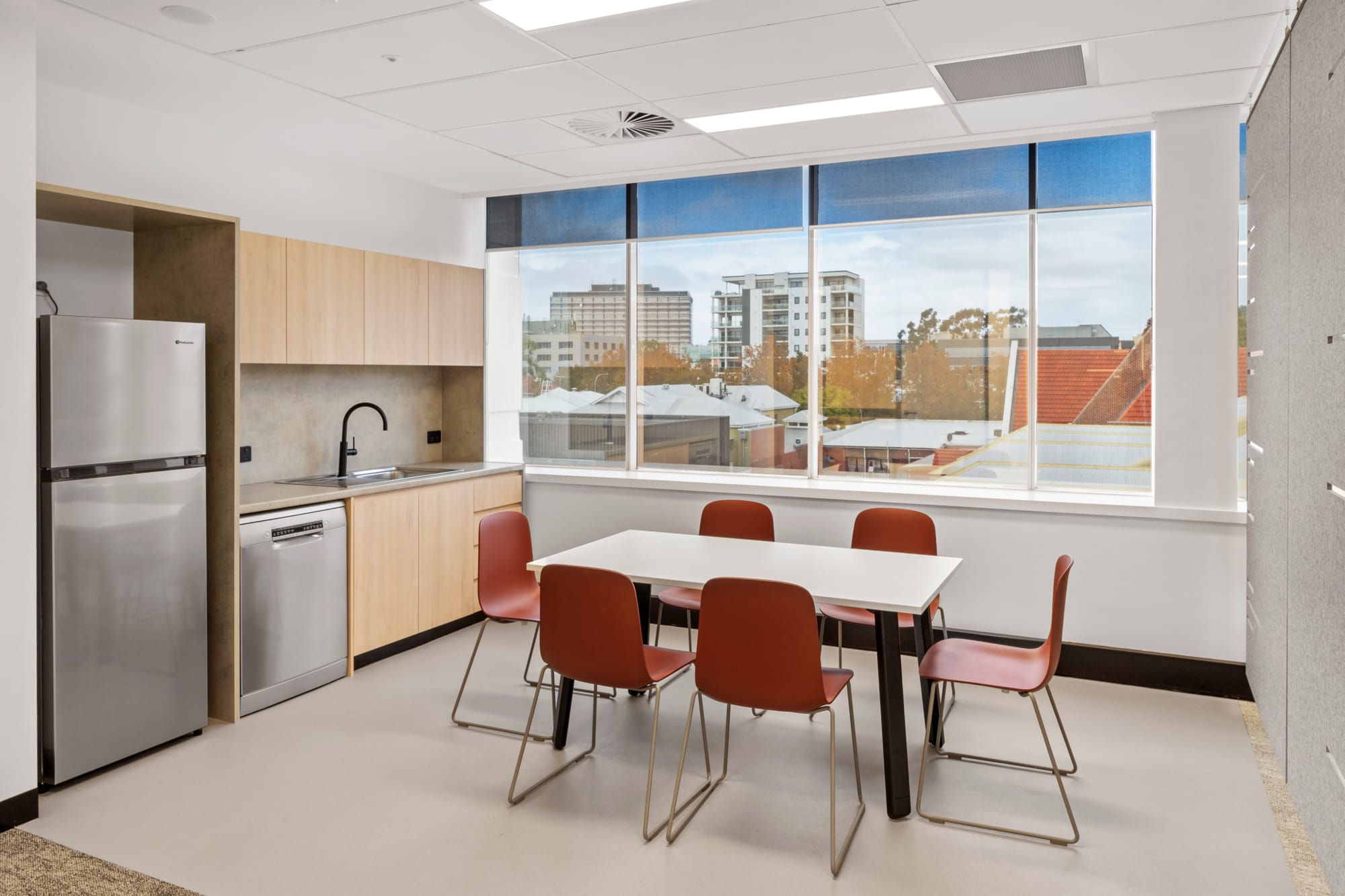 MDA National by Saru Design. Photography by Pete Battye. Office kitchen space with timber cabinetry, white flooring, white table with red chairs and city views out of a half-heigh window. 