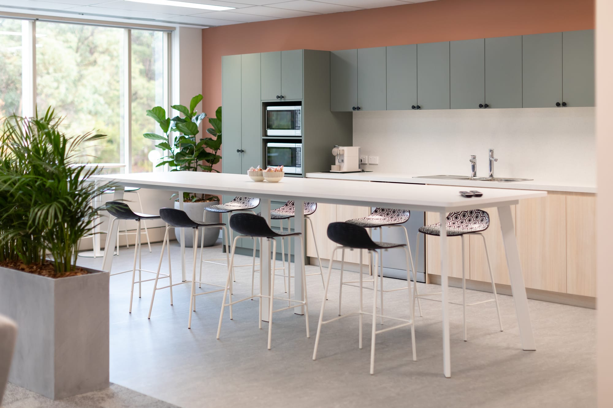 Telus Health by Saru Design. Photography by Ryan North. Office kitchen space with sage cabinets, terracotta walls, concrete floors and a long, white dining table. 