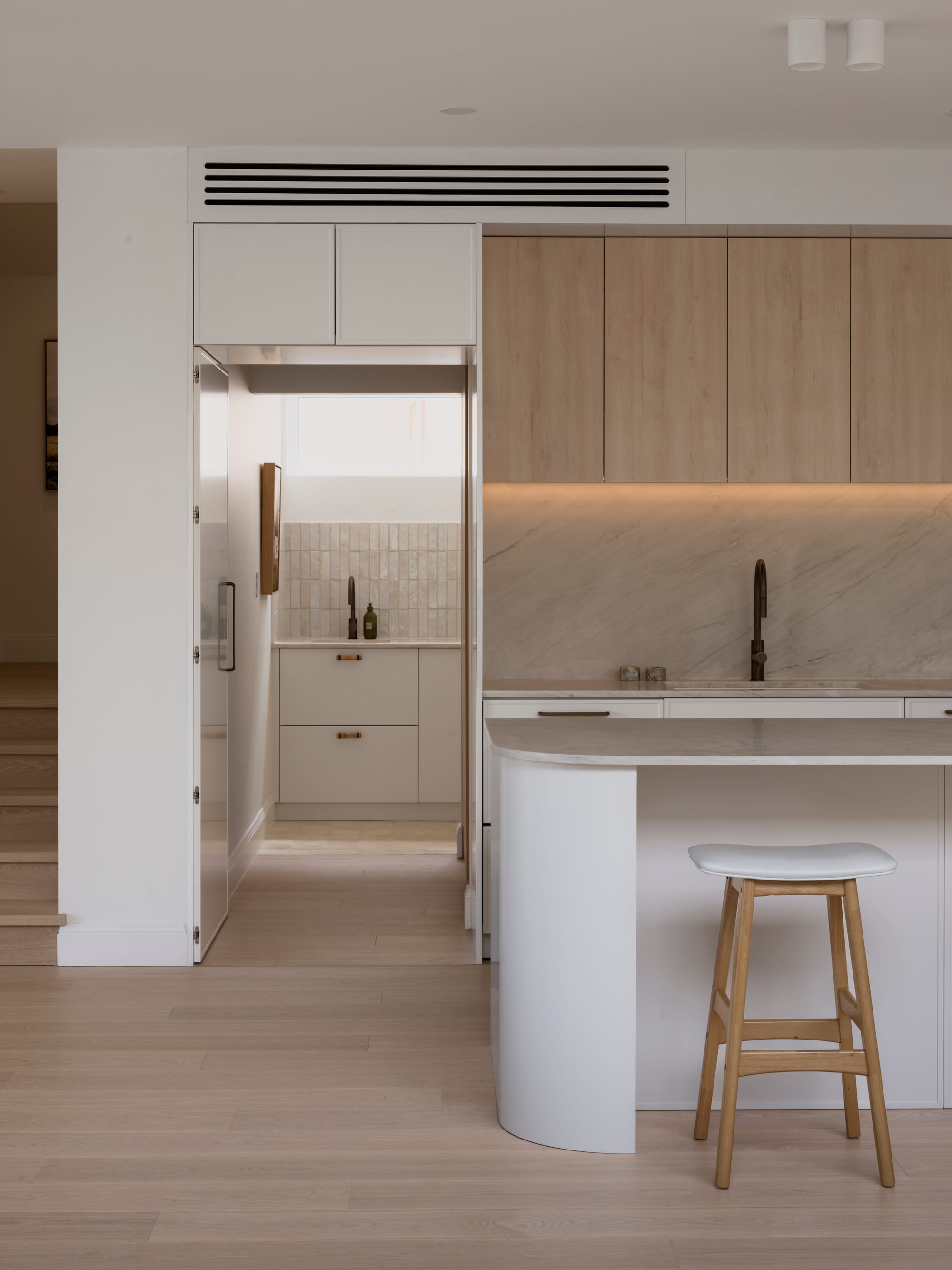 Sienna House by 868 Architects. Photography by Luke Butterly. Kitchen with white cabinetry, stone countertops and splashback, timber barstools and scullery with tiled stone splashback. 