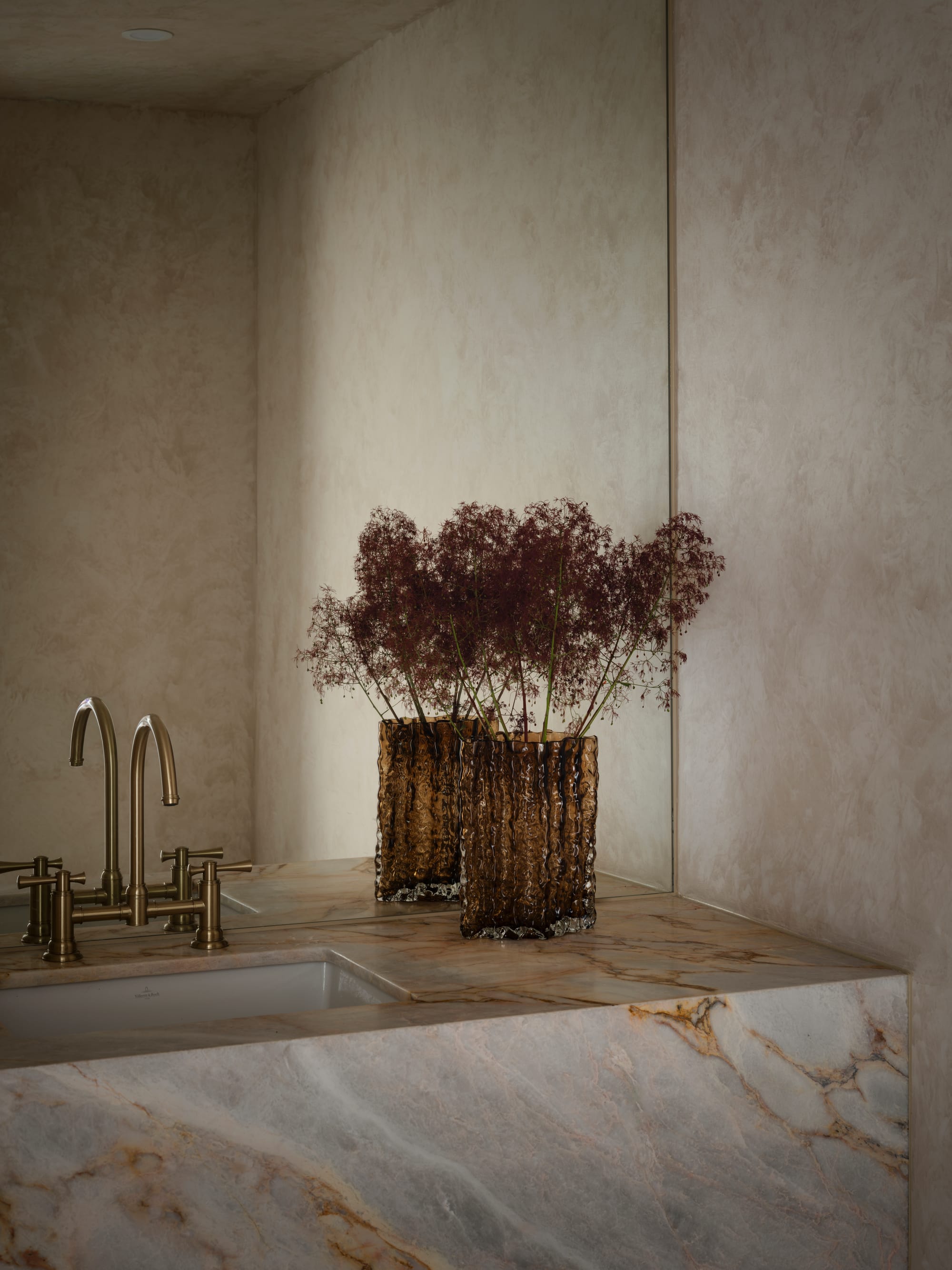 Sienna House by 868 Architects. Photography by Luke Butterly. Bathroom with heavily-veined stoned counters, lime-washed walls, mirrored splashback and dark glass vase.  