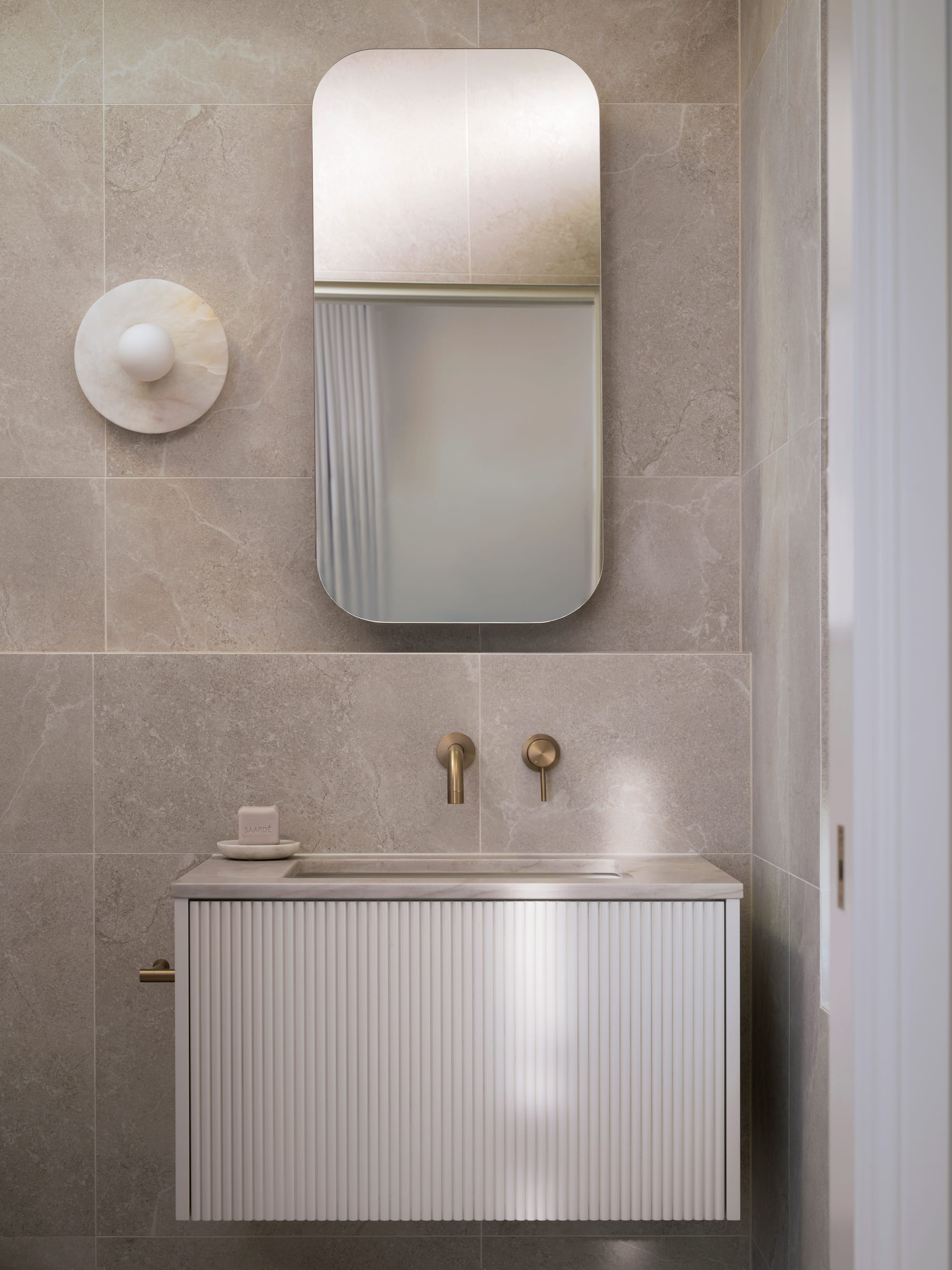 Sienna House by 868 Architects. Photography by Luke Butterly. Bathroom with white fluted timber cabinetry, stone countertop, tiled walls, and brass fixtures. 