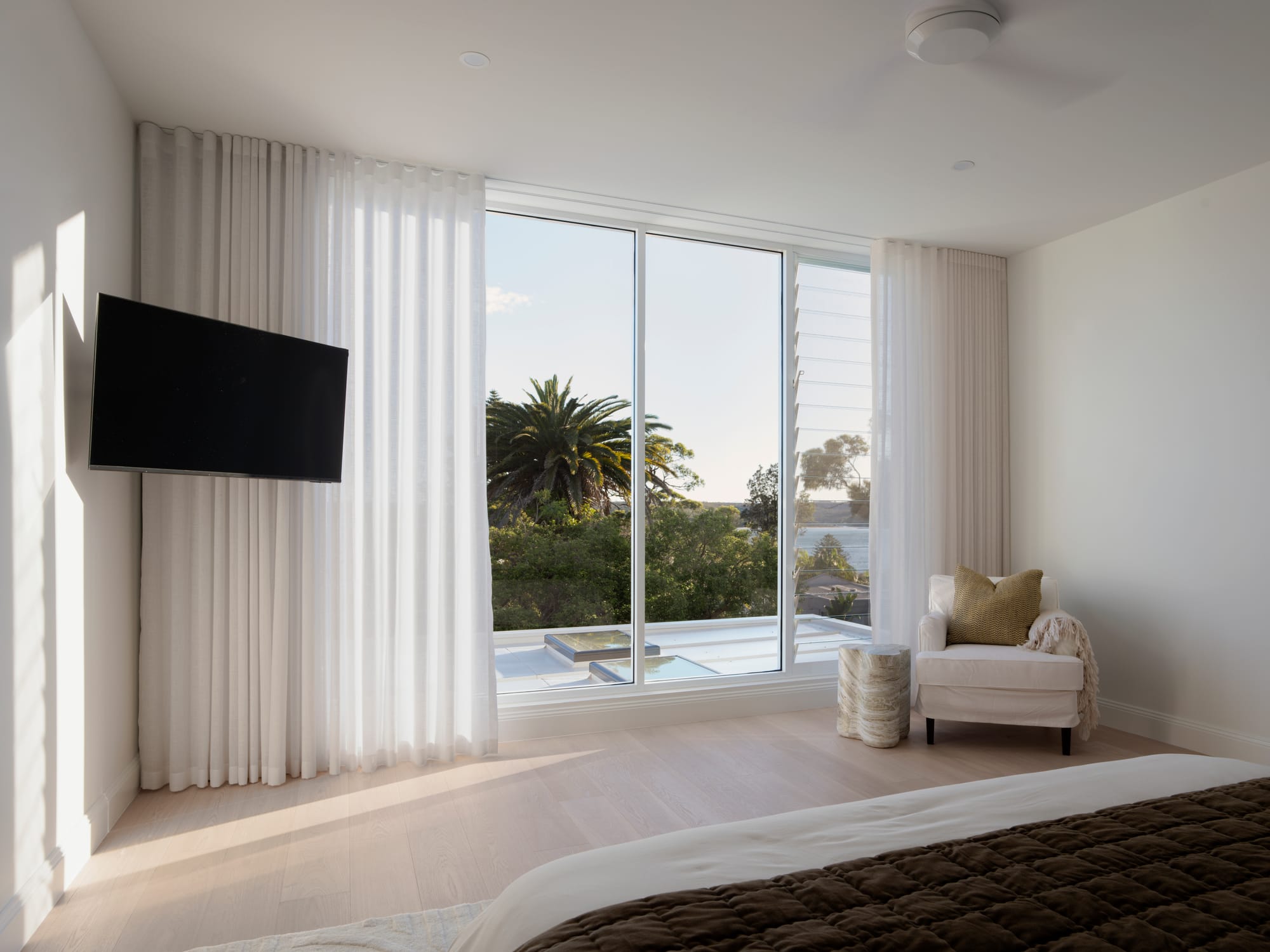 Sienna House by 868 Architects. Photography by Luke Butterly. Bedroom with white walls, timber floors and wall-mounted televisions, with full-height windows overlooking backyard. 