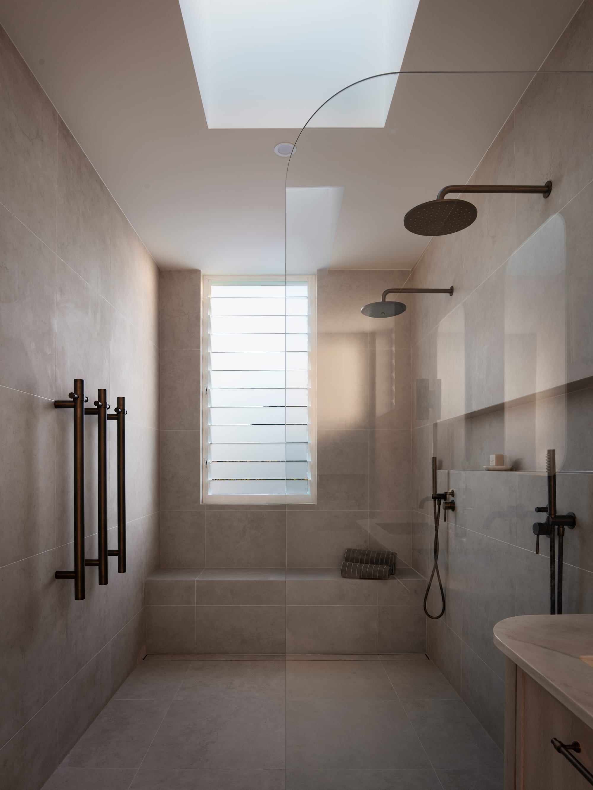 Sienna House by 868 Architects. Photography by Luke Butterly. Bathroom with tiled floors, walls and integrated seating, glass shower screen, brass fixtured and timber cabinetry. 