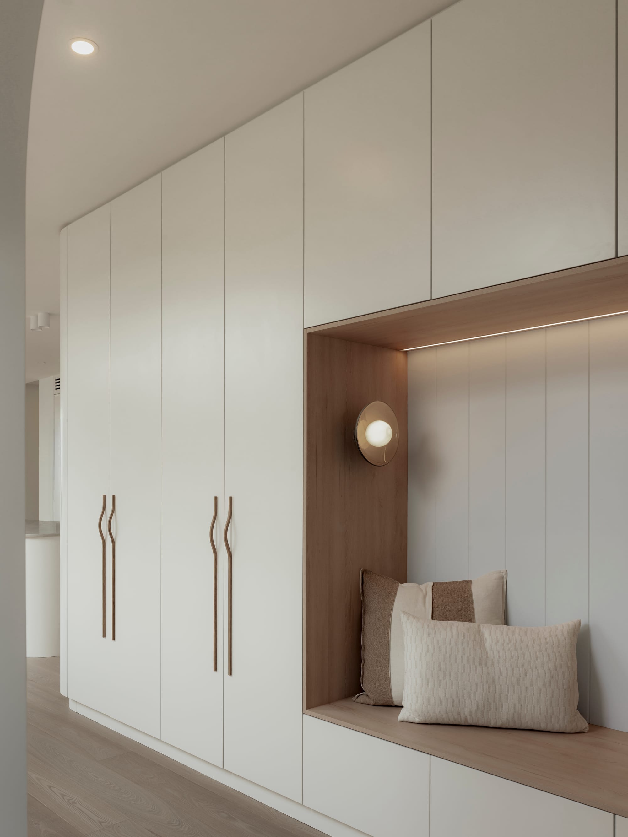 Sienna House by 868 Architects. Photography by Luke Butterly. Hallway with integrated white cabinetry, timber bench seat, timber flooring and beige throw cushions. 