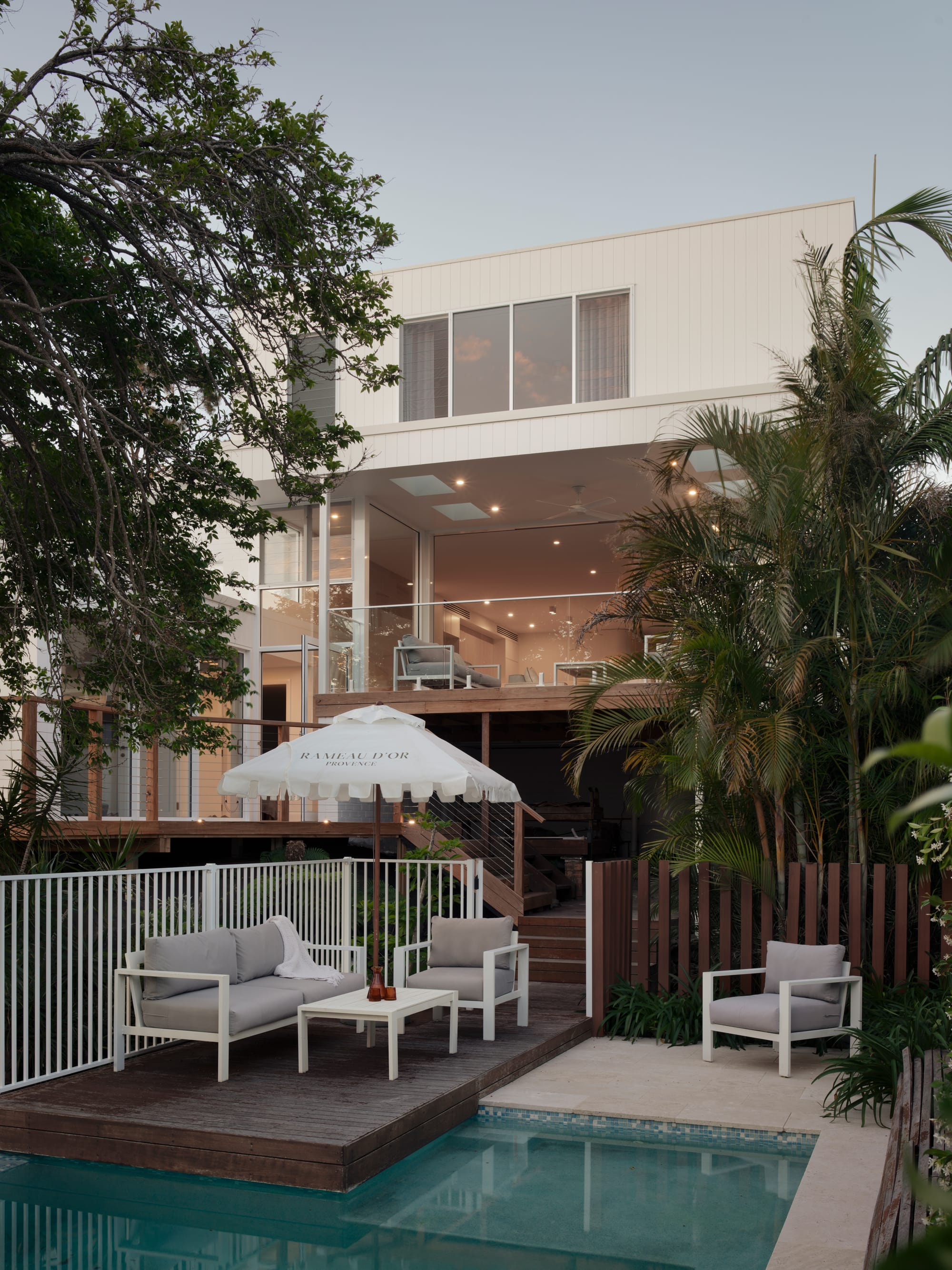 Sienna House by 868 Architects. Photography by Luke Butterly. Rear facade of home and backyard with timber decking, pool, dense foliage and white outdoor furniture. 