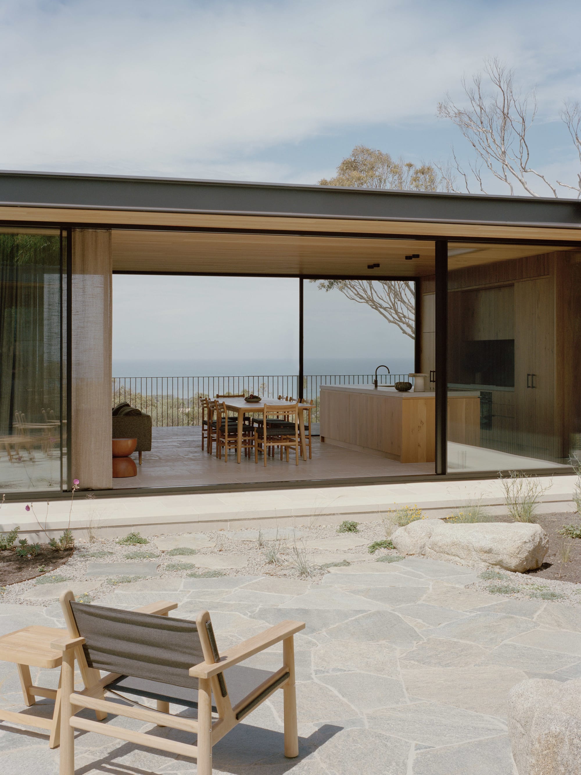 Anglesea House by Eckersley Architects. Photography by Tasha Tylee. Exterior courtyard with paved flooring and timber deck chair, overlooking open plan kitchen, dining and living spaces though full-height glass windows. 