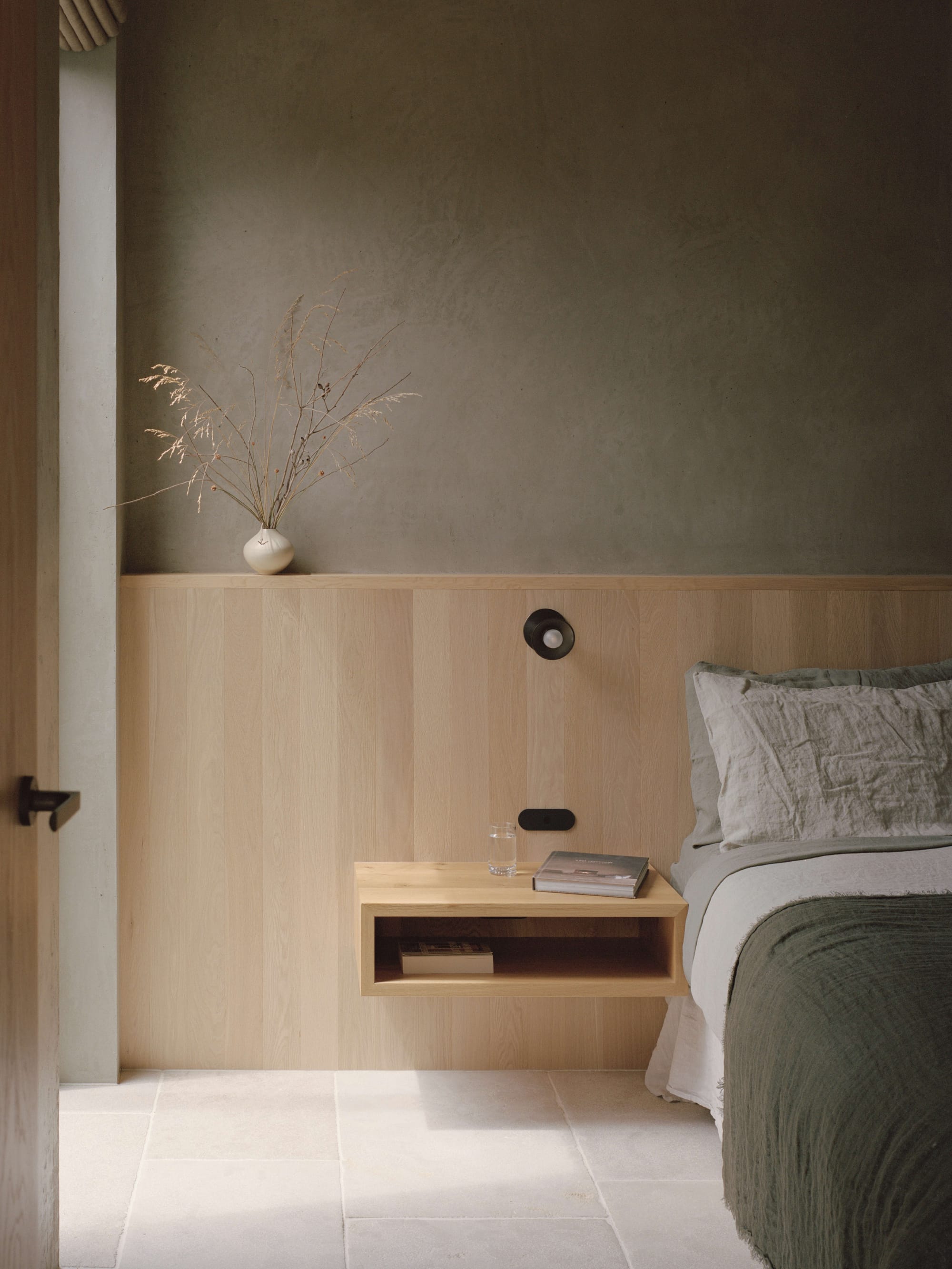 Anglesea House by Eckersley Architects. Photography by Tasha Tylee. Bedroom with olive and beige linen bedding, integrated timber headboard and side tables, lime-washed walls and stone flooring. 
