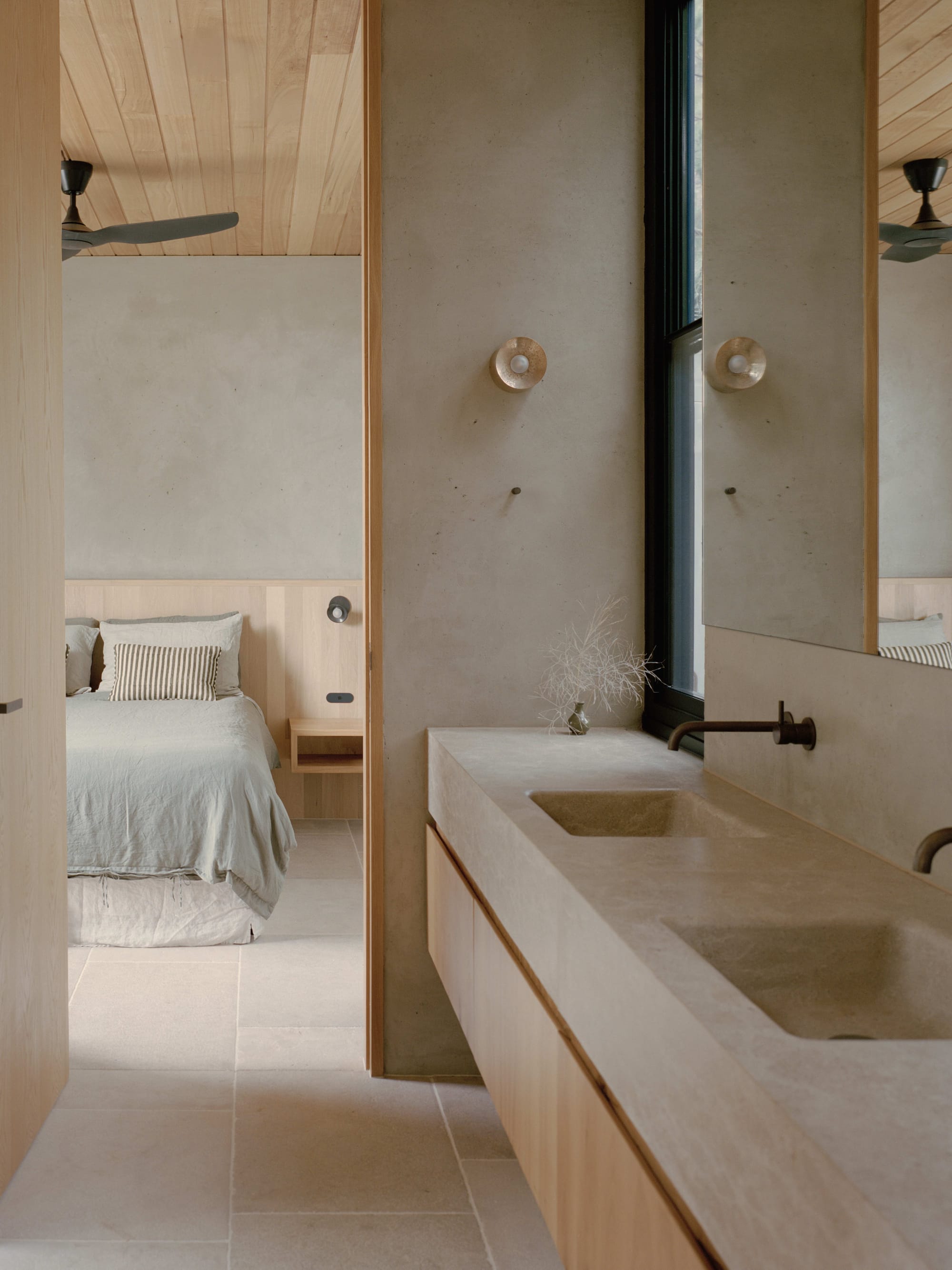 Anglesea House by Eckersley Architects. Photography by Tasha Tylee. Ensuite with stone countertops and integrated sinks, timber cabinetry and stone flooring, continuing through to bedroom with sky blue bed linen. 
