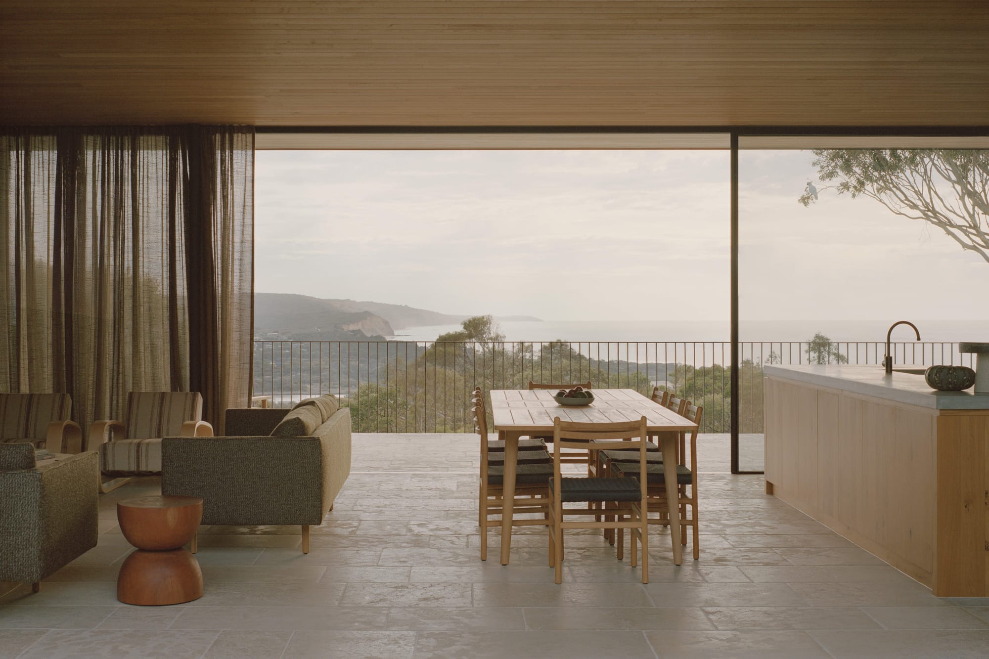 Anglesea House by Eckersley Architects. Photography by Tasha Tylee. Open plan living, kitchen and dining area with full-height glass windows overlooking balcony with timber decking, black balustrade and panoramic views of coast and bush. 