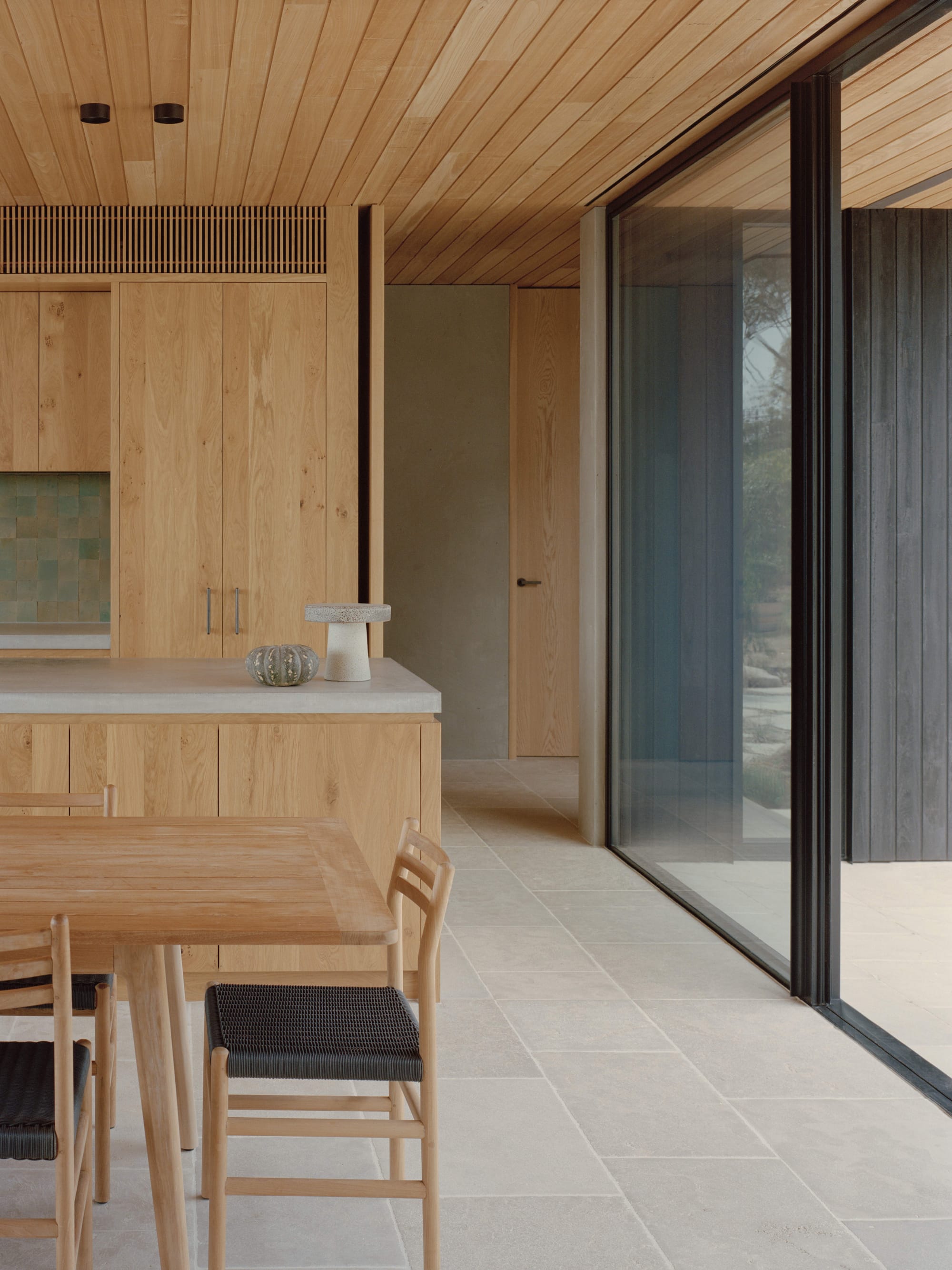 Anglesea House by Eckersley Architects. Photography by Tasha Tylee. Open plan kitchen and dining room with timber cabinetry, stone countertop, stone tiled floor, timber dining table and full-height glass windows. 