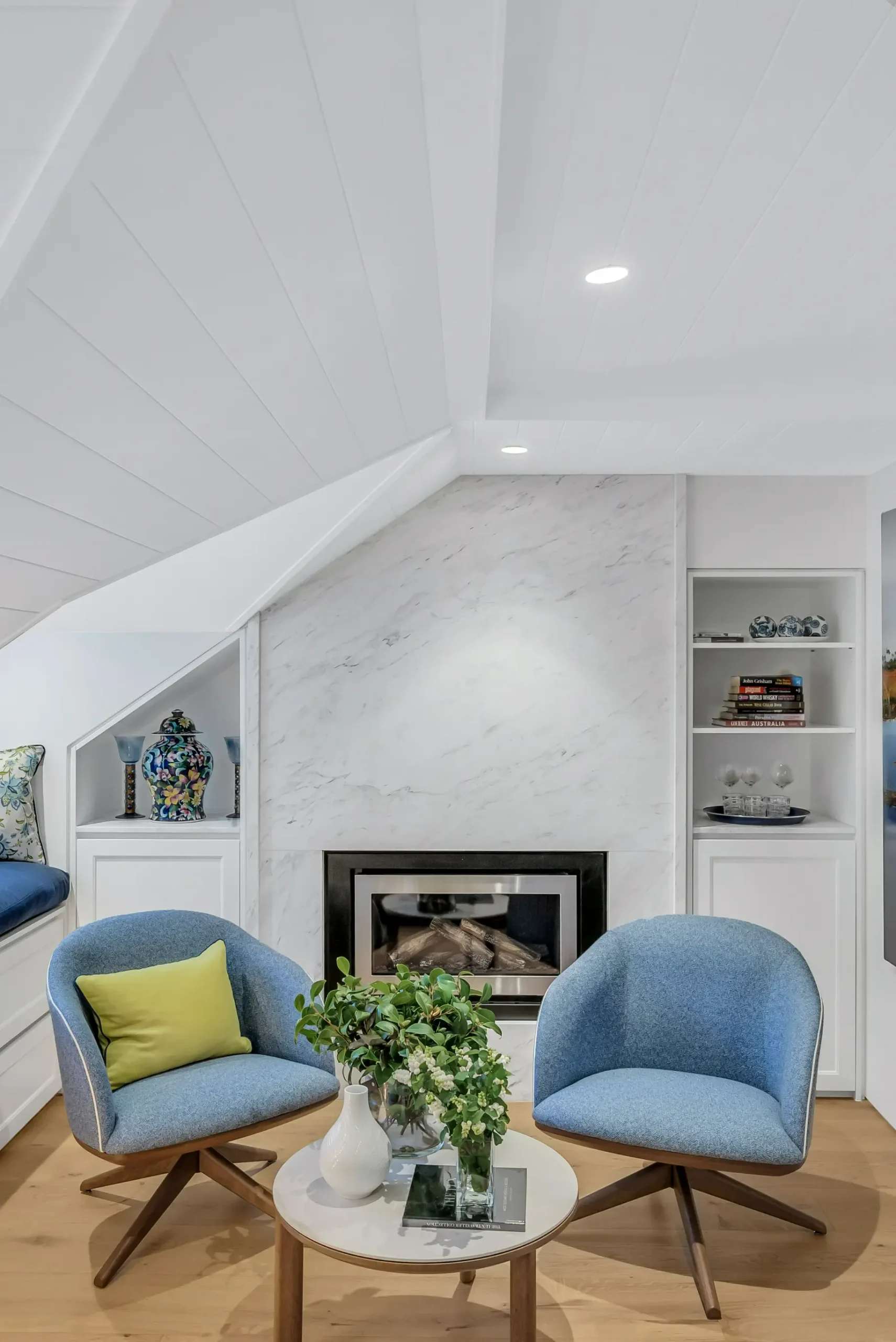 Cozy Elegance by Emotive Designs. Two blue armchairs in sitting room with timber floors, marble fireplace and angled white ceiling. 