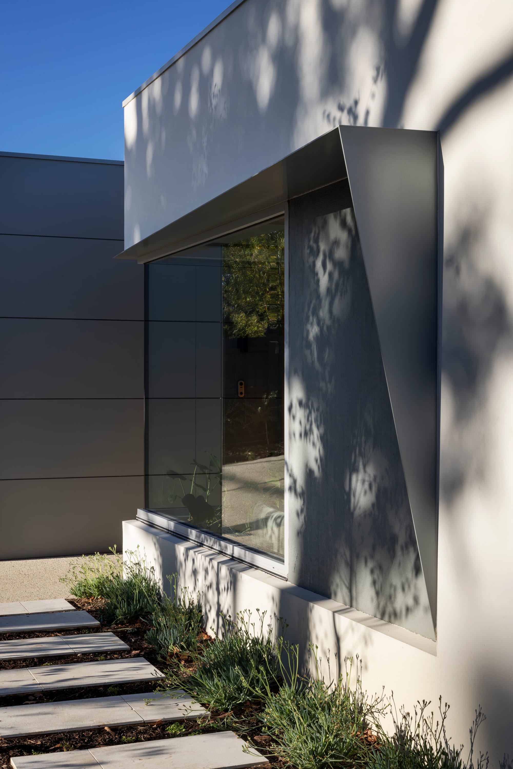 Earth + Sky House by Kirsten Johnstone Architecture. Photography by Tatjana Plitt. Front facade of home with grey finish, abstract grey shade covering over window, and pavers through garden