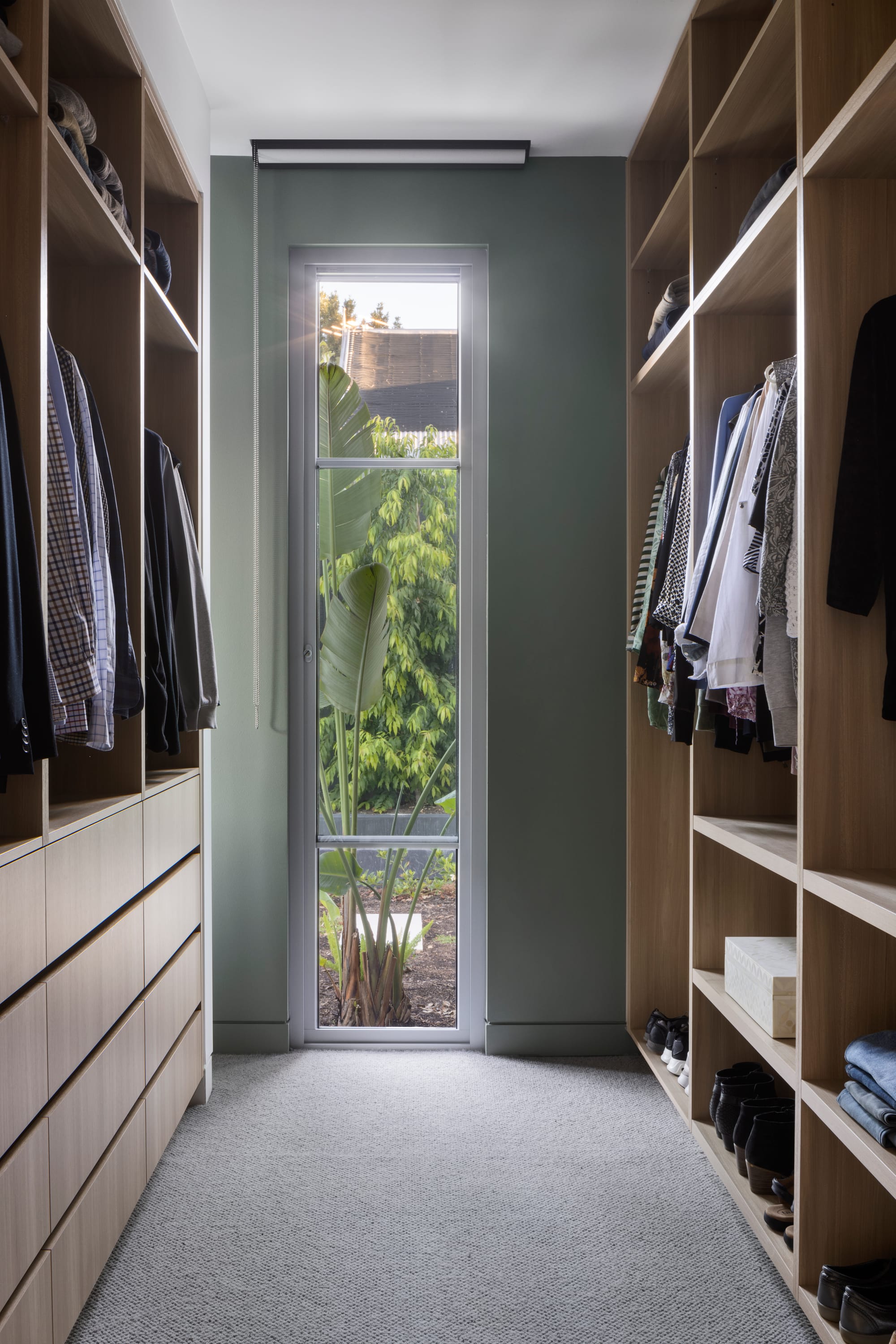 Earth + Sky House by Kirsten Johnstone Architecture. Photography by Tatjana Plitt. Walk in robe with grey flooring, integrated timber cabinetry, sage walls and thin window overlooking garden. 