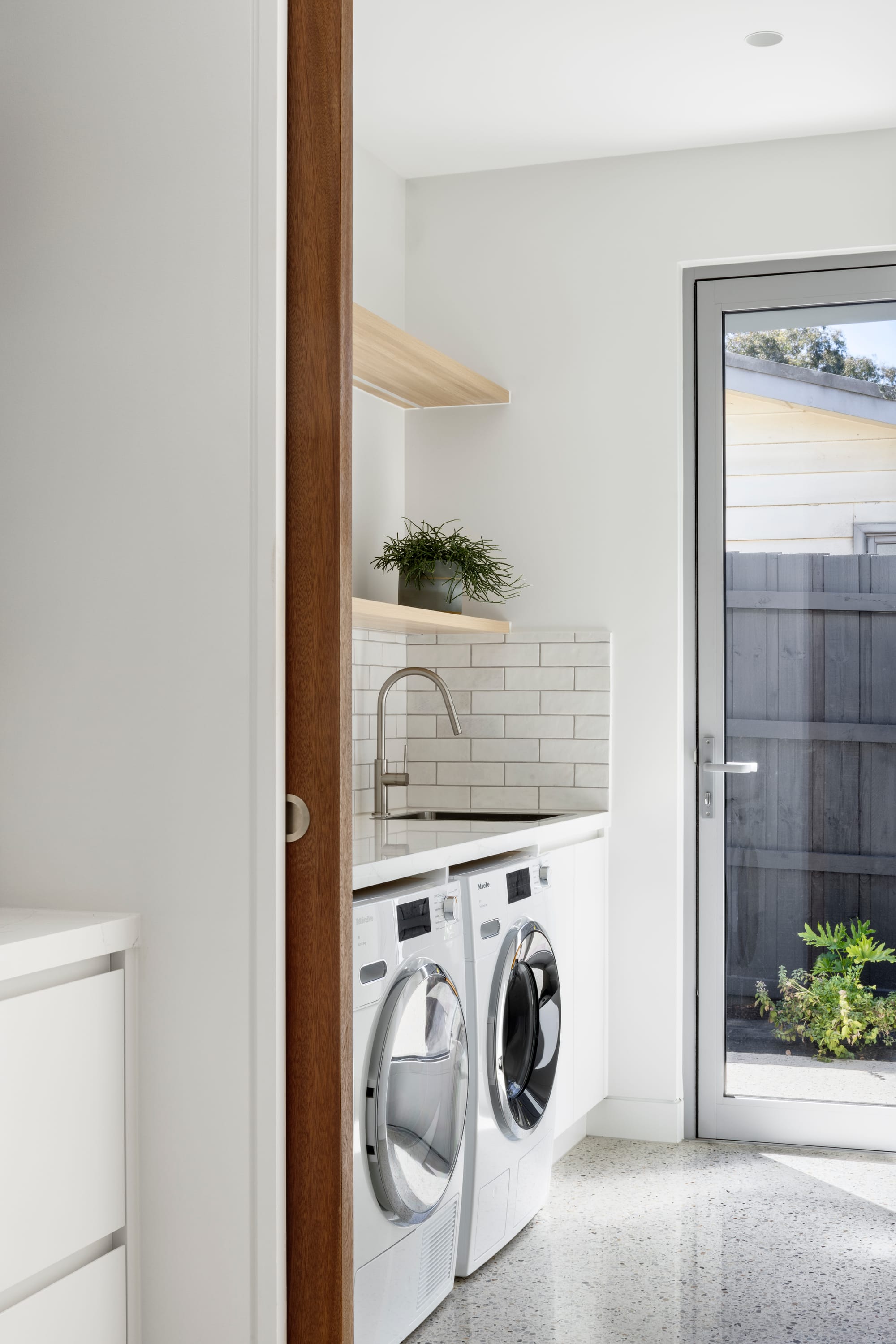 Earth + Sky House by Kirsten Johnstone Architecture. Photography by Tatjana Plitt. Laundry with polised concrete floors, white cabinetry and appliances, subway tile splashback and glass door opening onto garden. 