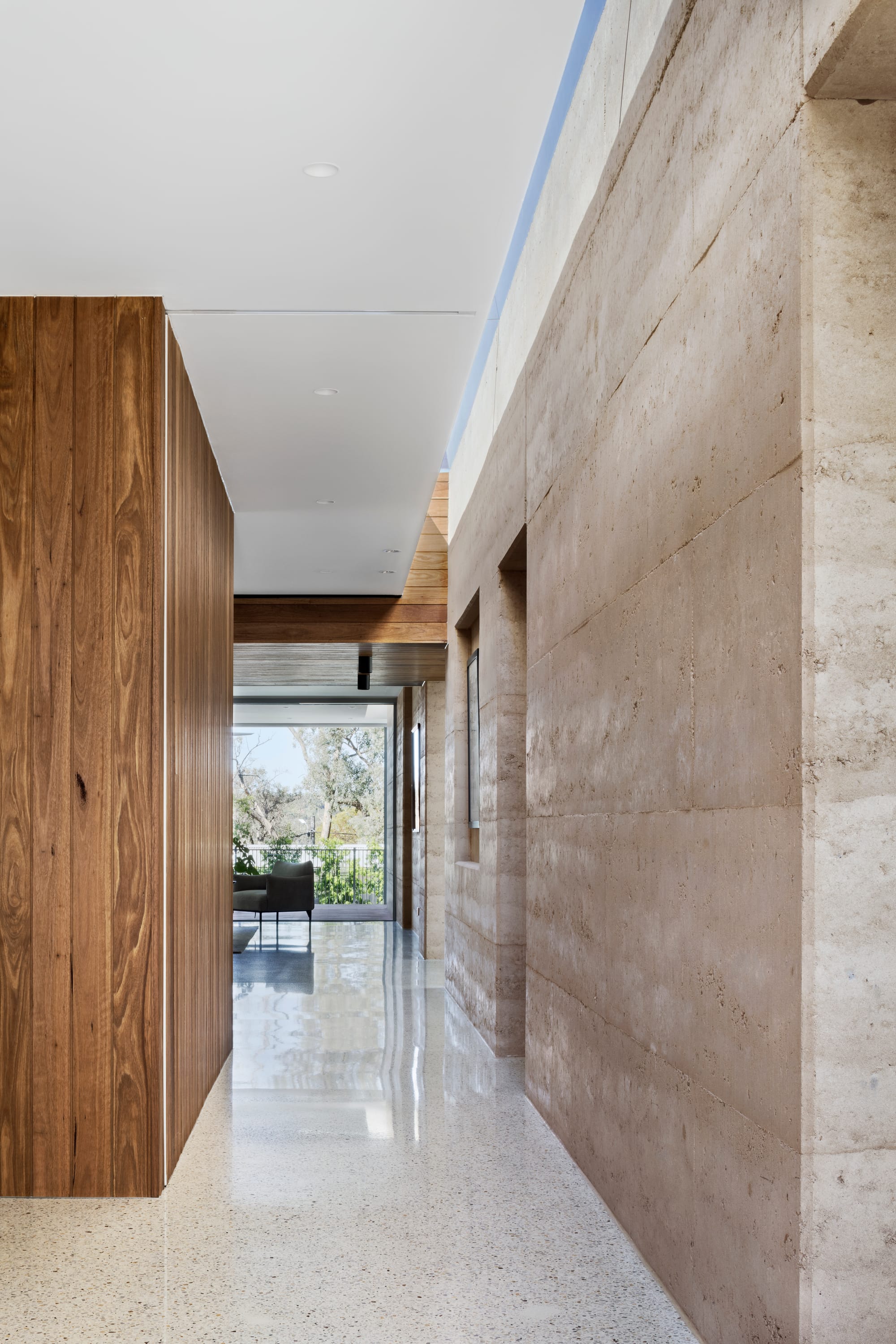 Earth + Sky House by Kirsten Johnstone Architecture. Photography by Tatjana Plitt. Hallway with polished concrete flooring, timber clad walls on left and rammed earth walls on right, leading to living space and outdoor timber deck. 